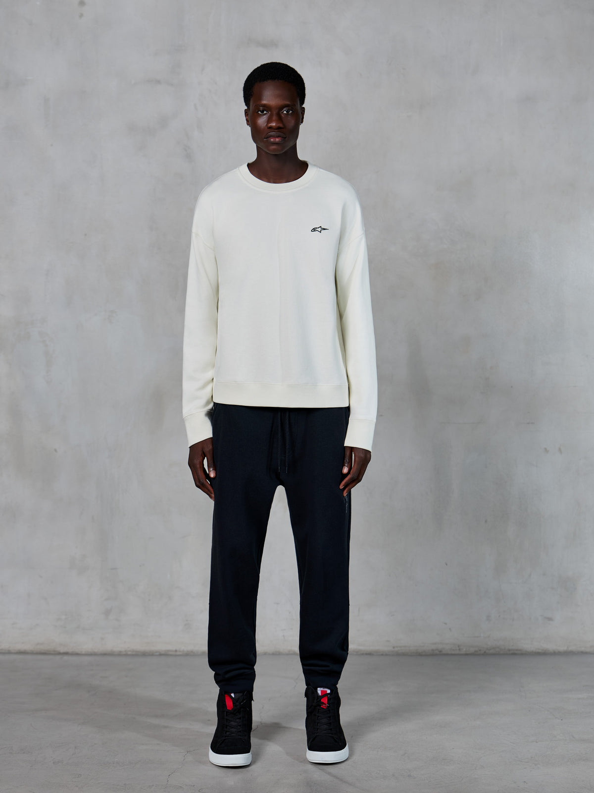 Alpinestars Essentials Ageless Crew, sweatshirt de gola redonda, Off White com pequeno logótipo de estrela preto, usada por um modelo com calças jogger pretas, streetwear casual inspirado no desporto motorizado