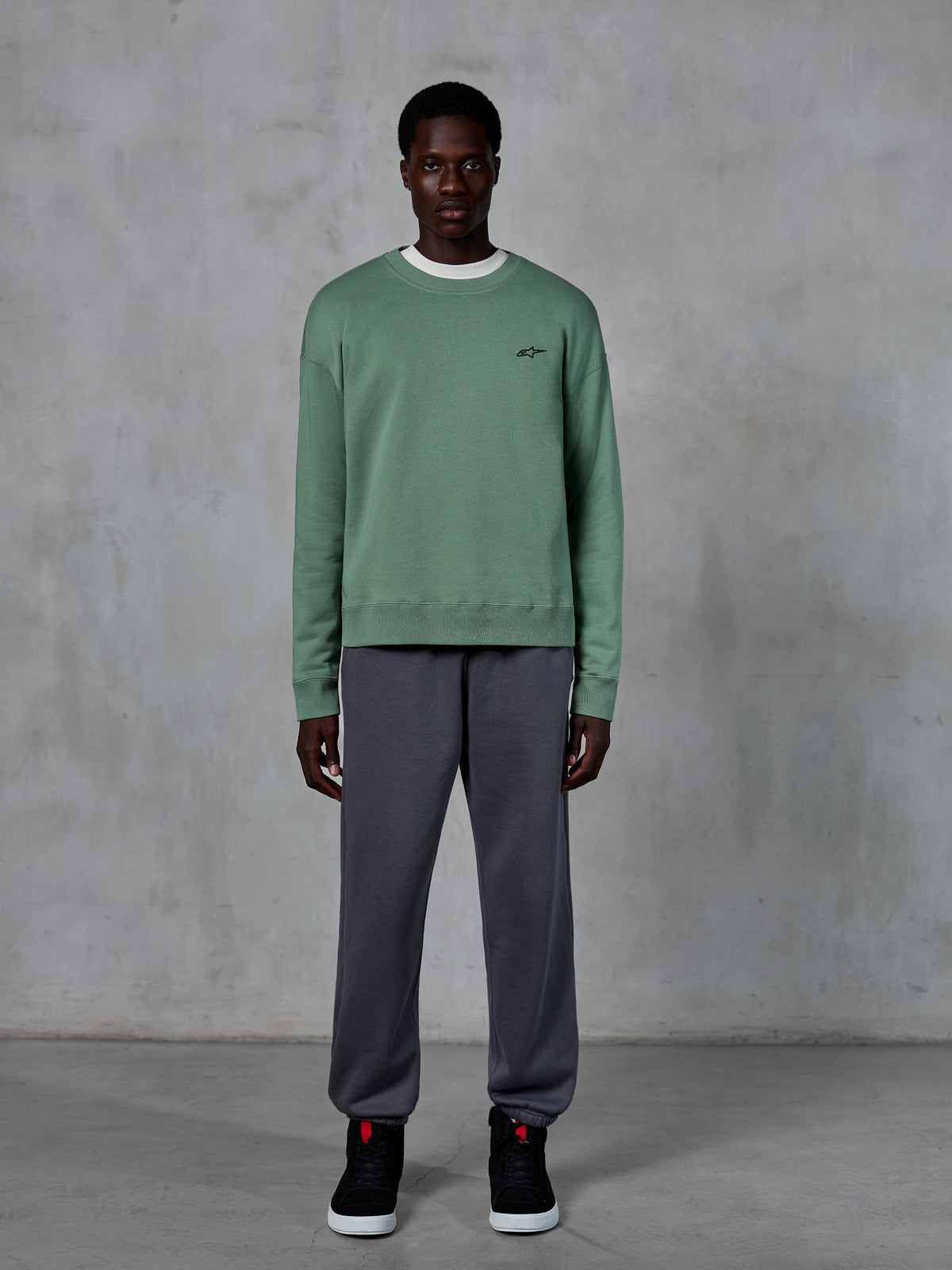 Alpinestars Essentials Ageless Crew, sweatshirt de gola redonda em verde Cactus, com um pequeno logótipo preto no peito, usada por um modelo com calças de fato de treino cinzentas e sapatilhas pretas, streetwear casual inspirado no desporto motorizado