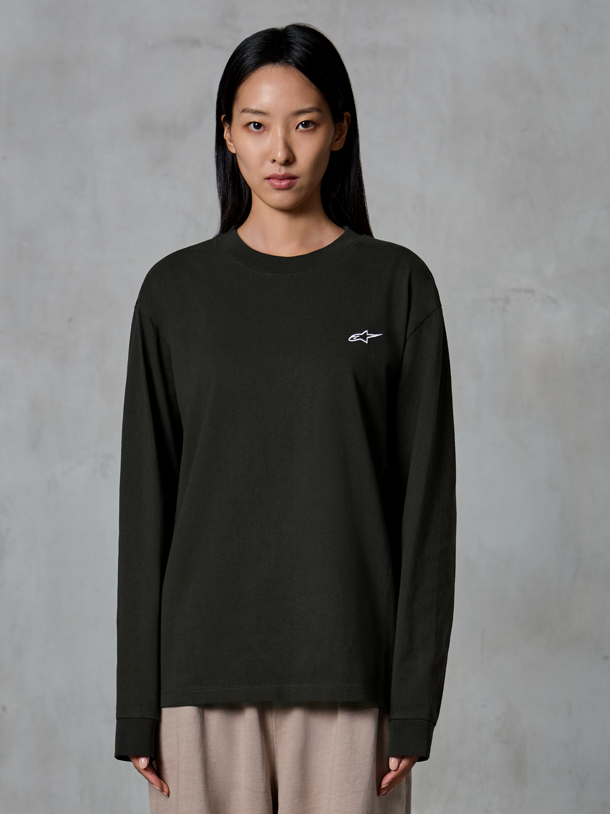 Model wearing Alpinestars Essentials Ageless LS Tee, Long Sleeve T-Shirt, Off Black, featuring a small white Alpinestars logo on the chest, paired with beige trousers, casual lifestyle streetwear