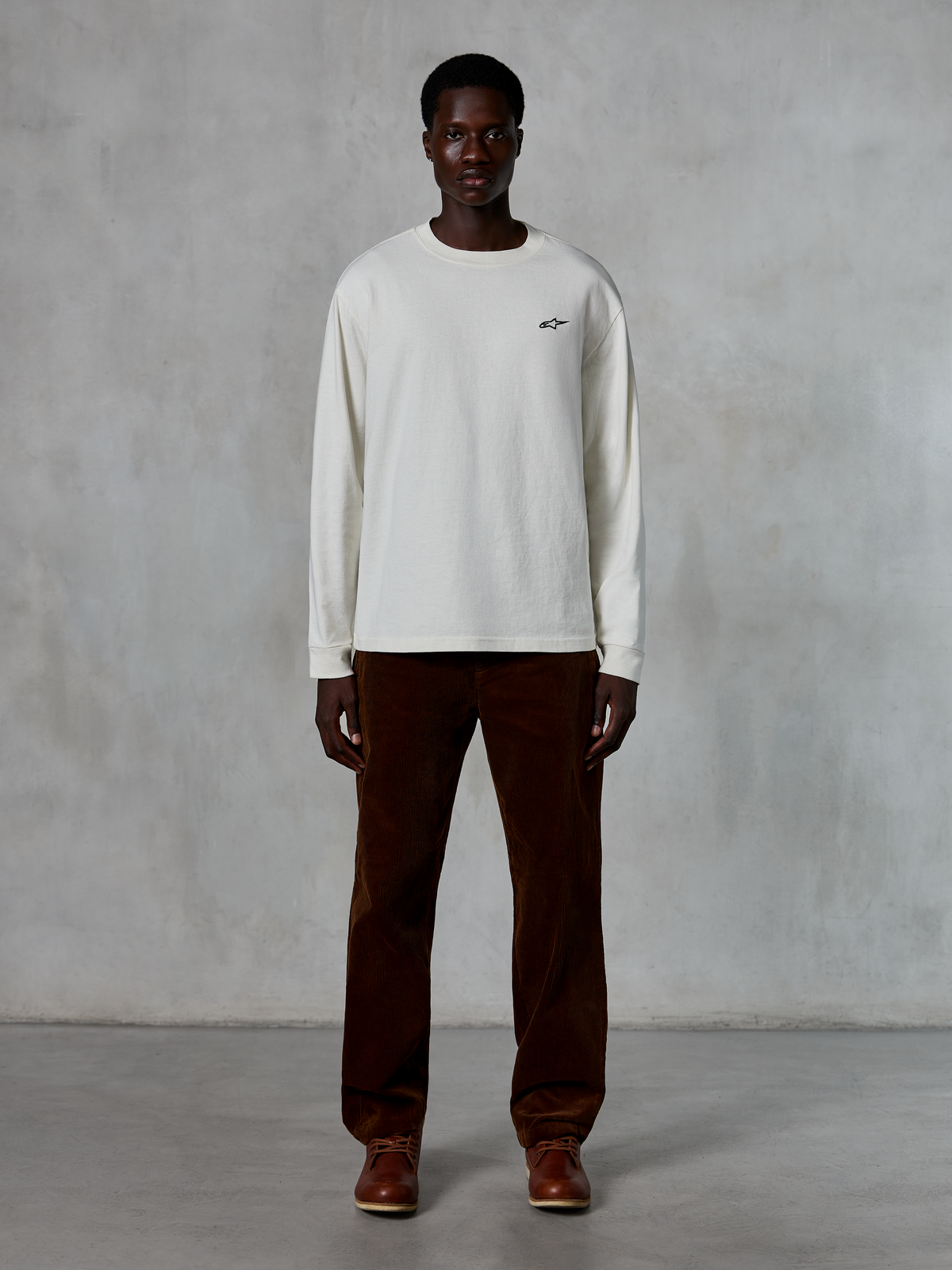 Alpinestars Essentials Ageless LS Tee, Long-Sleeve T-Shirt, Off-White, worn by model with brown corduroy pants and brown leather boots, featuring a small black logo on the chest, relaxed contemporary fit, casual lifestyle streetwear