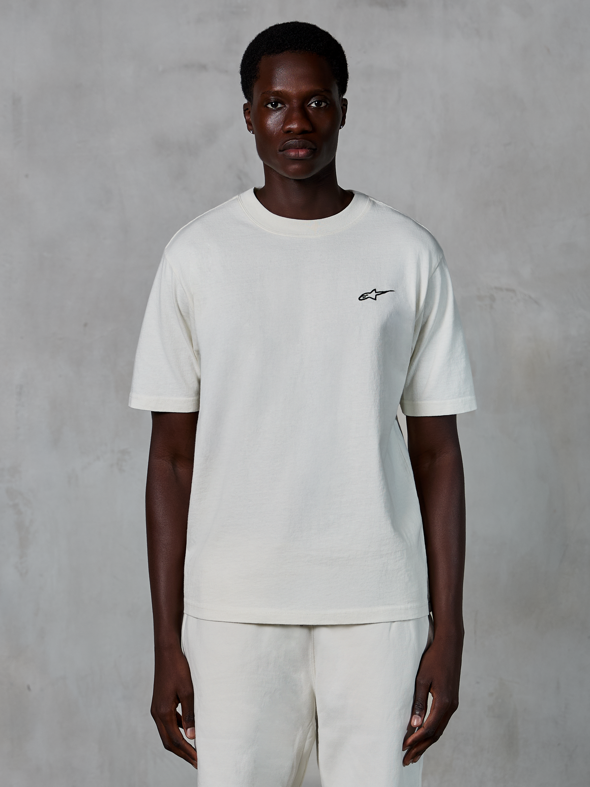 Alpinestars Essentials Ageless SS Tee, t-shirt de manga curta, Off White, usada por um modelo masculino, com um pequeno logótipo Alpinestars preto no peito, corte relaxado, jersey de algodão orgânico, streetwear casual de desportos motorizados