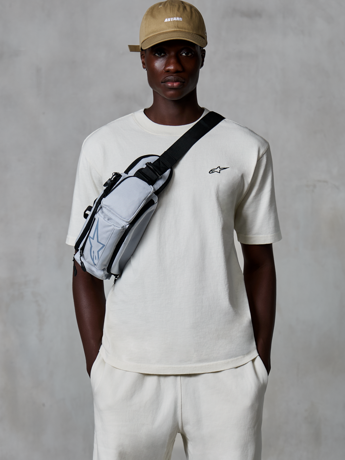Alpinestars Essentials Ageless SS Tee, t-shirt de manga curta, Off White, usada com calções a condizer e um boné bege, com um pequeno logótipo preto no peito e uma bolsa de tiracolo cinzenta, streetwear casual inspirado no desporto motorizado