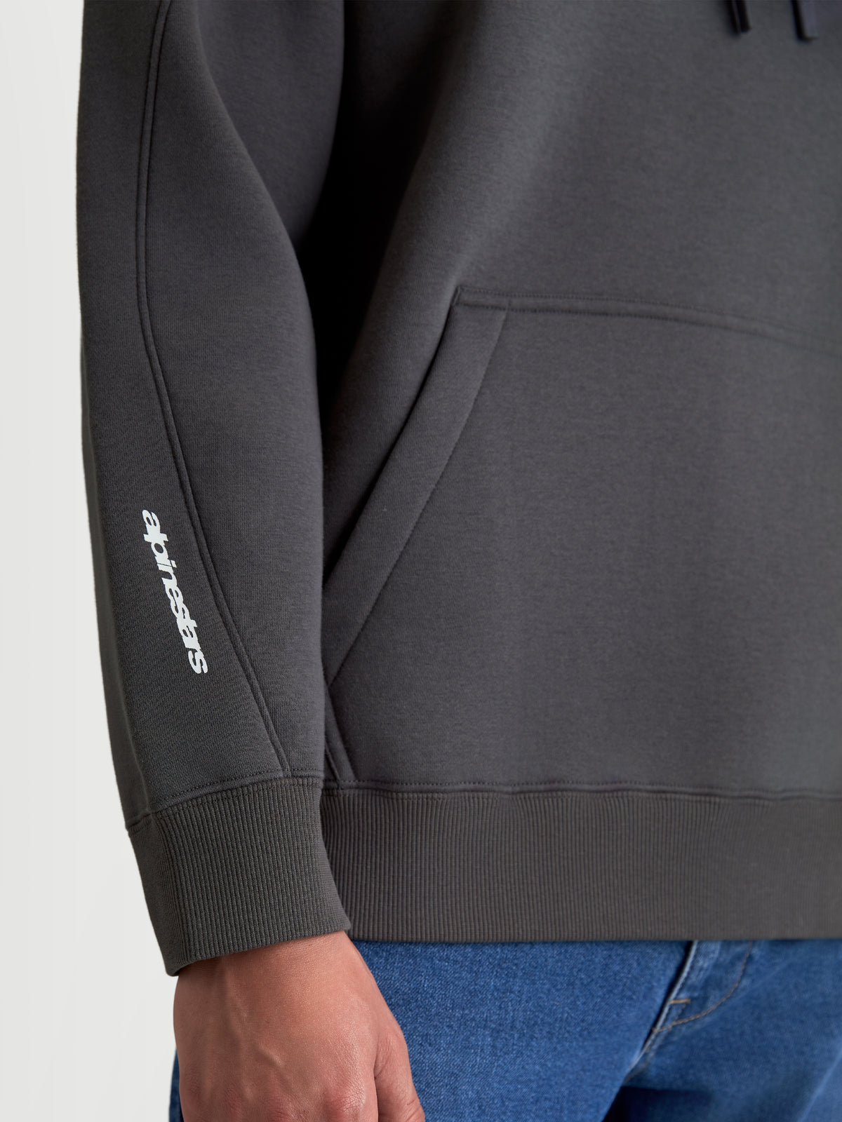 Alpinestars Prelude Oversized Hoodie, camisola com capuz cinzenta em fleece, vista de detalhe mostrando as costuras da manga, o punho canelado e o logótipo Alpinestars branco no braço, sportswear com ajuste descontraído e bolso canguru.