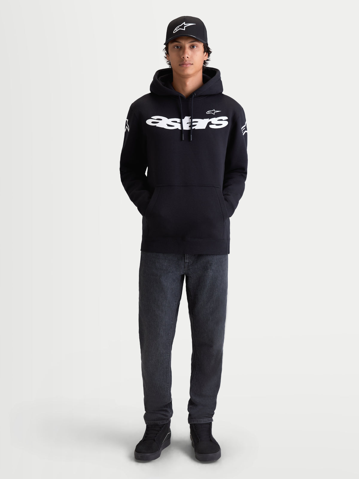 Alpinestars Camisola Com Capuz Velocifier, camisola com capuz para homem, preta, com logótipo Astars branco na frente e nos ombros, sportswear casual para uso fora da moto, modelo a usar boné e calças de ganga.