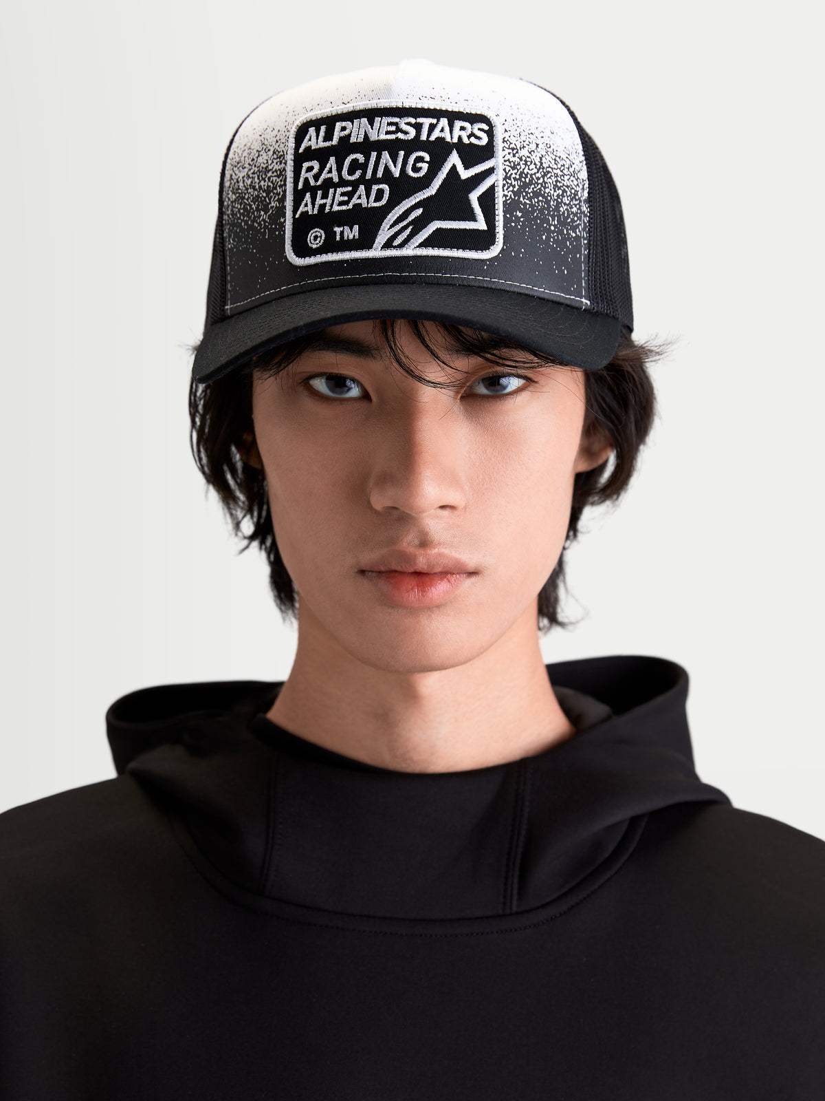 Alpinestars boné trucker Gravel, boné sportswear lifestyle, preto e branco, vista frontal no modelo com pala curva, malha traseira respirável e patch tecido Racing Ahead com detalhe de textura de cascalho.
