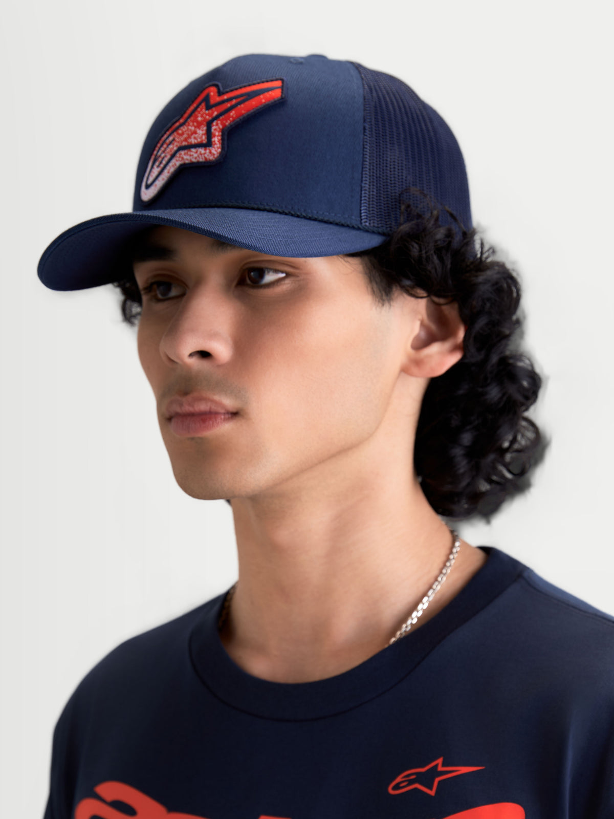 Alpinestars Boné Trucker Meteorite, boné sportswear com pala curva e coroa estruturada, azul marinho com logótipo Astars vermelho em textura de cascalho, traseira em malha, vista num modelo.