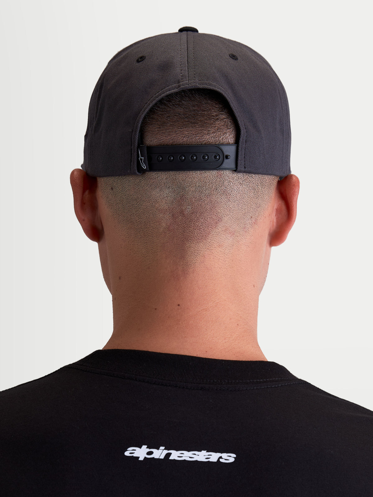 Alpinestars Boné Stadium Snapback, vista traseira num modelo mostrando o fecho snapback regulável e a coroa estruturada, cinzento escuro e preto, sportswear em sarja de algodão durável.