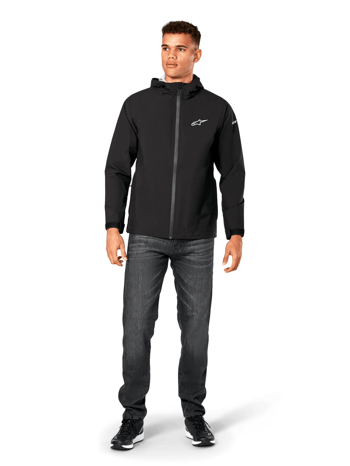 Alpinestars Kascade Rain Jacket, casaco impermeável para homem, preto, usado com calças de ganga cinzento-escuras e sapatilhas pretas, com capuz, fecho frontal revestido e logótipo Alpinestars branco no peito, streetwear casual inspirado no desporto motorizado