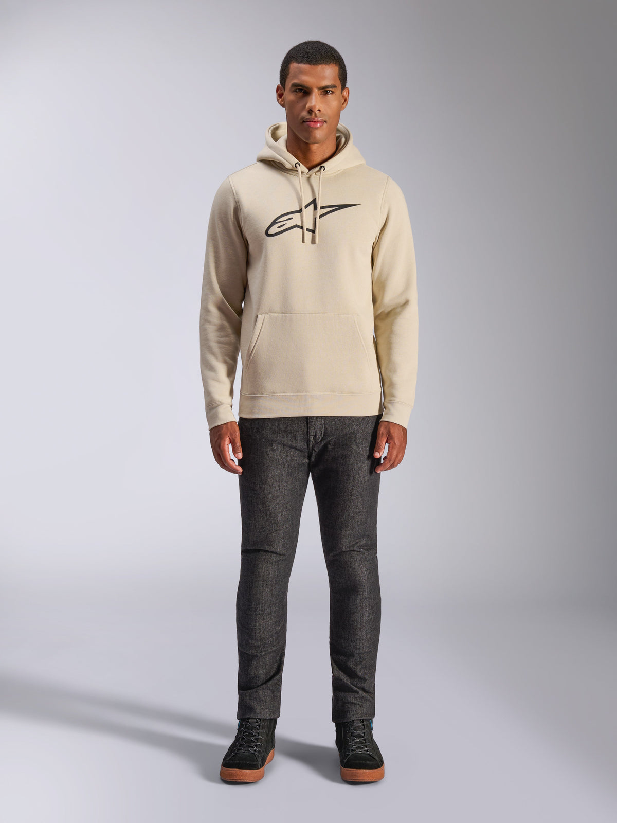 Alpinestars Ageless V3 Hoodie, casual sportswear, Off White with black embroidered logo, worn by a man with dark grey pants and black sneakers, featuring a drawstring hood and kangaroo pocket, modern motorsport-inspired design