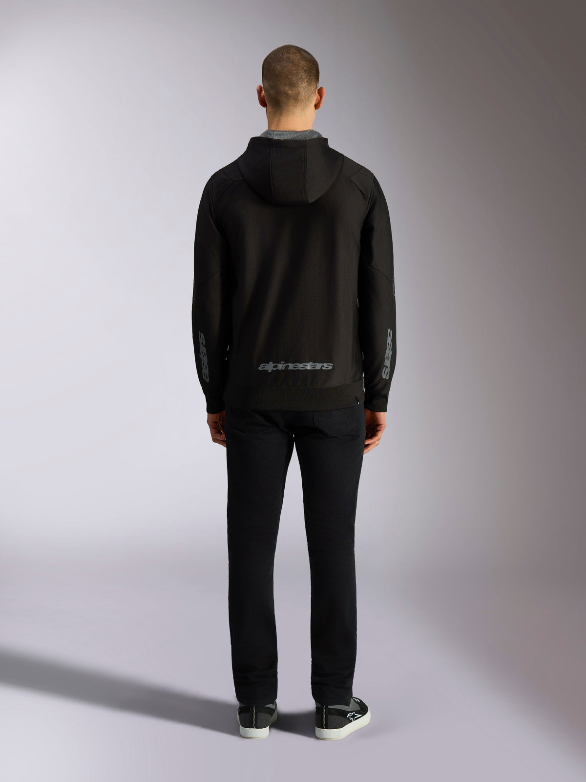Vista traseira do Hoodie Alpinestars Sessions V3 em Preto e Cinzento, com o logótipo Alpinestars a cinzento na parte inferior das costas e logótipos astars nos antebraços, usado por um modelo com calças pretas, streetwear casual inspirado no desporto motorizado