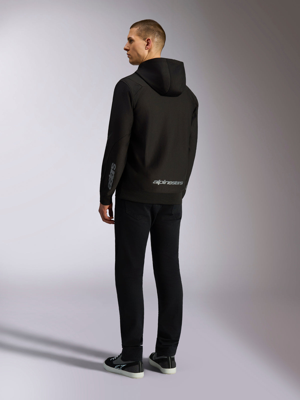 Alpinestars Sessions V3 Hoodie, streetwear casual preto, vista traseira num modelo, com a marca nominativa Alpinestars a cinzento na parte inferior das costas e o logótipo Astars na manga, design com capuz e fecho frontal com estilo moderno inspirado no desporto motorizado