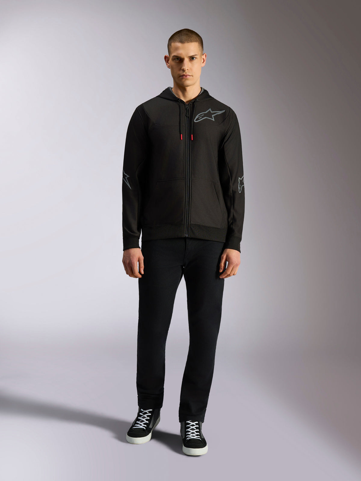 Camisola com capuz Alpinestars Sessions V3, hoodie preto com fecho integral, logótipo Astars cinzento no peito, gráficos nos cotovelos e antebraços, detalhes vermelhos nos cordões, usado por modelo masculino com calças pretas e sapatilhas, vestuário casual de desportos motorizados