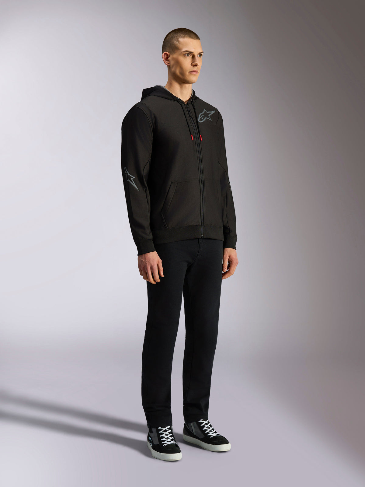 Hoodie Alpinestars Sessions V3, streetwear casual de motorsport, hoodie preto com fecho de correr, logótipo de estrela cinzento no peito e na manga, cordões com pontas vermelhas, punhos e bainha canelados, vestido por modelo com calças pretas e sapatilhas.