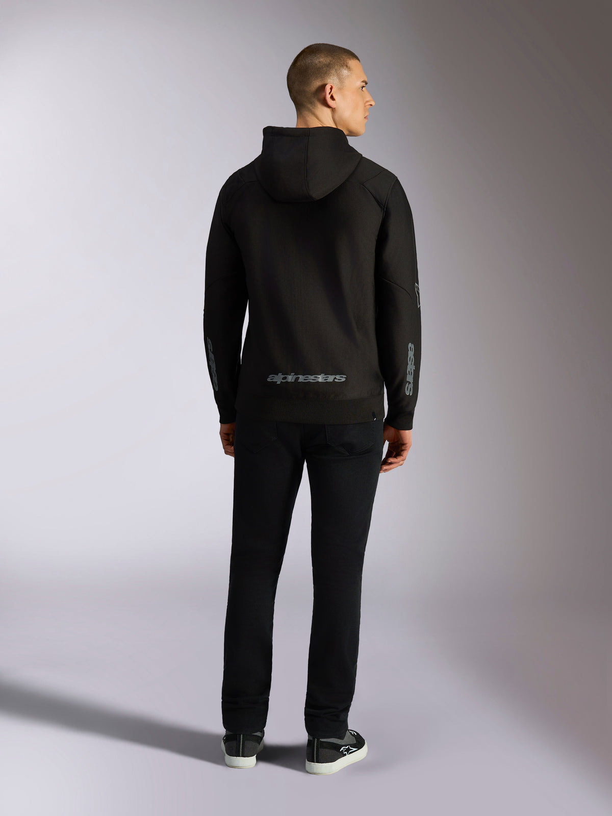 Alpinestars Sessions V3 Hoodie, streetwear casual, preto com a marca nominativa Alpinestars em cinzento na parte inferior das costas e logótipo 'astars' nas mangas, vista traseira de modelo masculino a usar o hoodie e calças pretas, design moderno inspirado no desporto motorizado