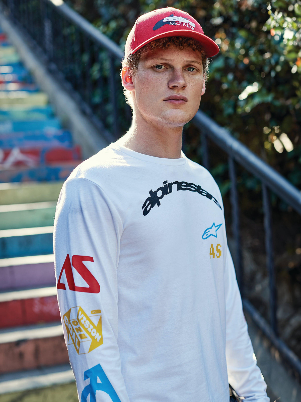 Boné Alpinestars Profile, vermelho, usado por um jovem, aba curva com coroa estruturada e logótipo bordado, combinado com uma t-shirt branca de manga comprida com grafismos, streetwear casual de motorsport