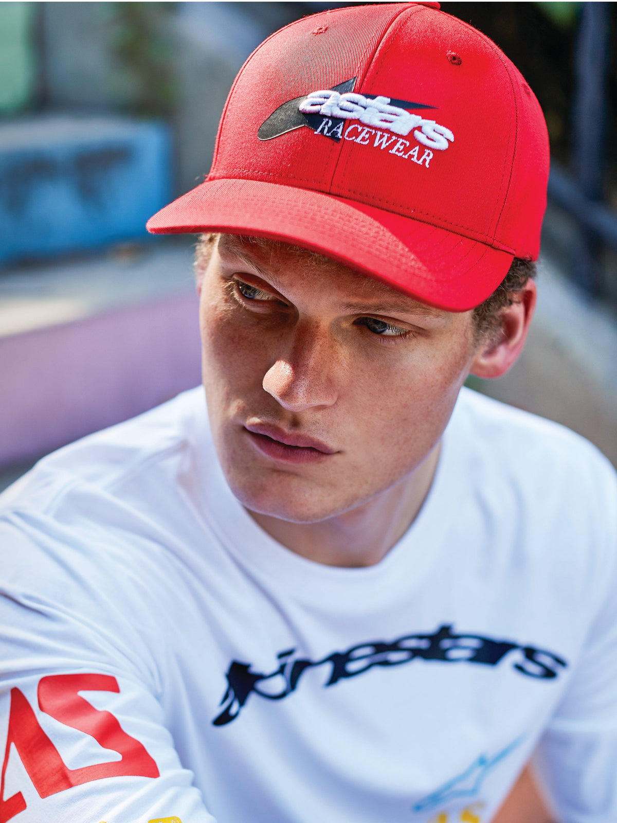 Homem a usar o Boné Profile da Alpinestars, boné snapback vermelho de aba curva com bordado frontal 'astars RACEWEAR' em branco e preto, combinado com uma t-shirt gráfica branca de manga comprida, estilo casual motorsport