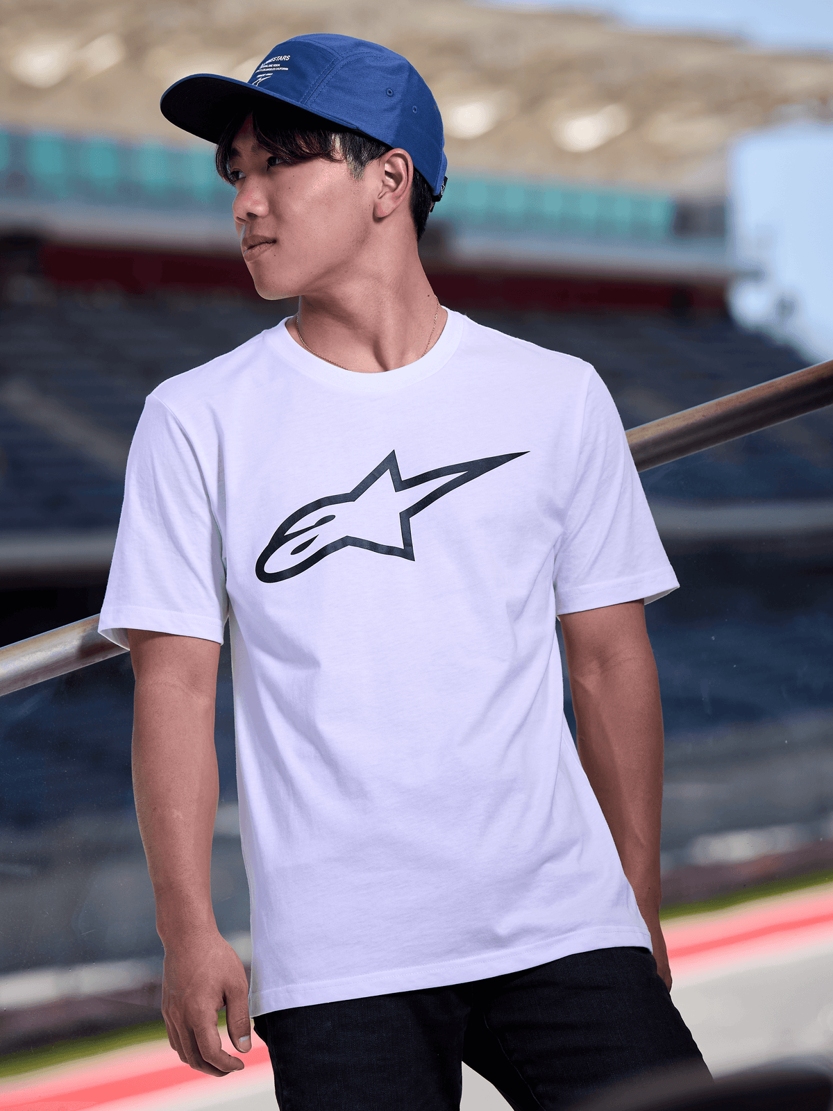 Homem a usar o Boné Alpinestars Peasy em azul, boné jockey de 5 painéis com pala plana e impressão gráfica do logótipo a branco, combinado com uma T-shirt branca com logótipo Alpinestars, streetwear casual inspirado no desporto motorizado