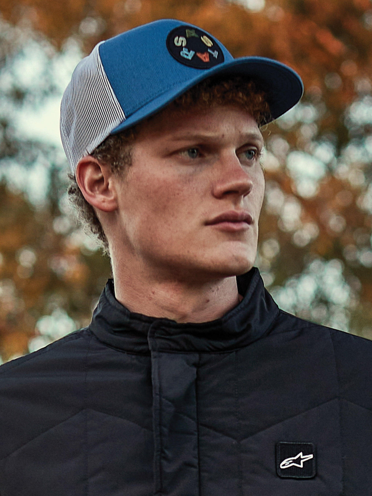 Man wearing Alpinestars Gambit Trucker Hat, Blue and White mesh-back cap with a circular Astars logo patch, paired with a black Alpinestars quilted jacket, casual motorsport-inspired lifestyle portrait with autumn background