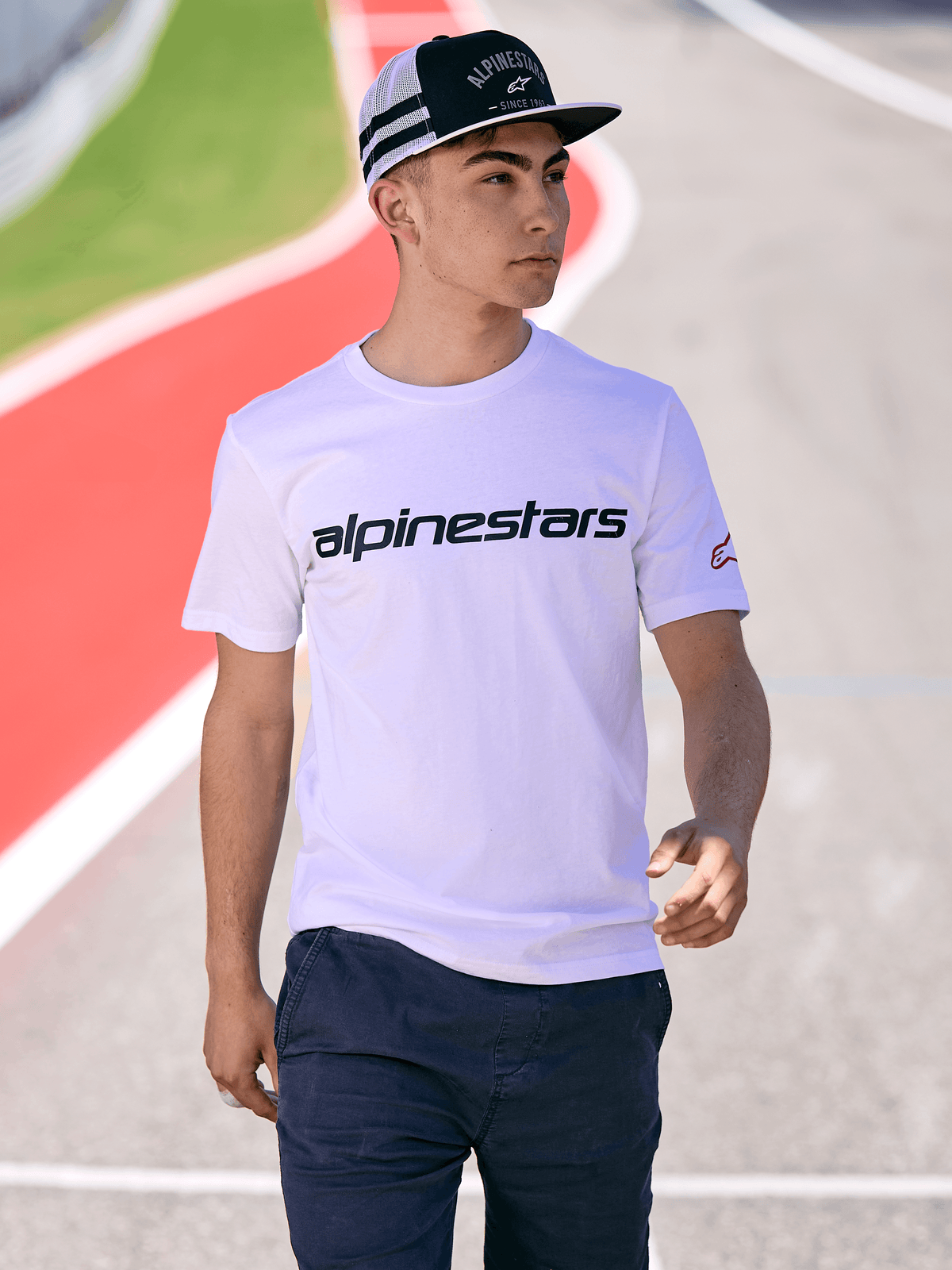 Homem a usar o boné Alpinestars Backline Trucker em preto e branco, combinado com uma t-shirt branca com o logótipo Alpinestars e calças azul-escuro, estilo streetwear casual de desportos motorizados num fundo de pista ao ar livre