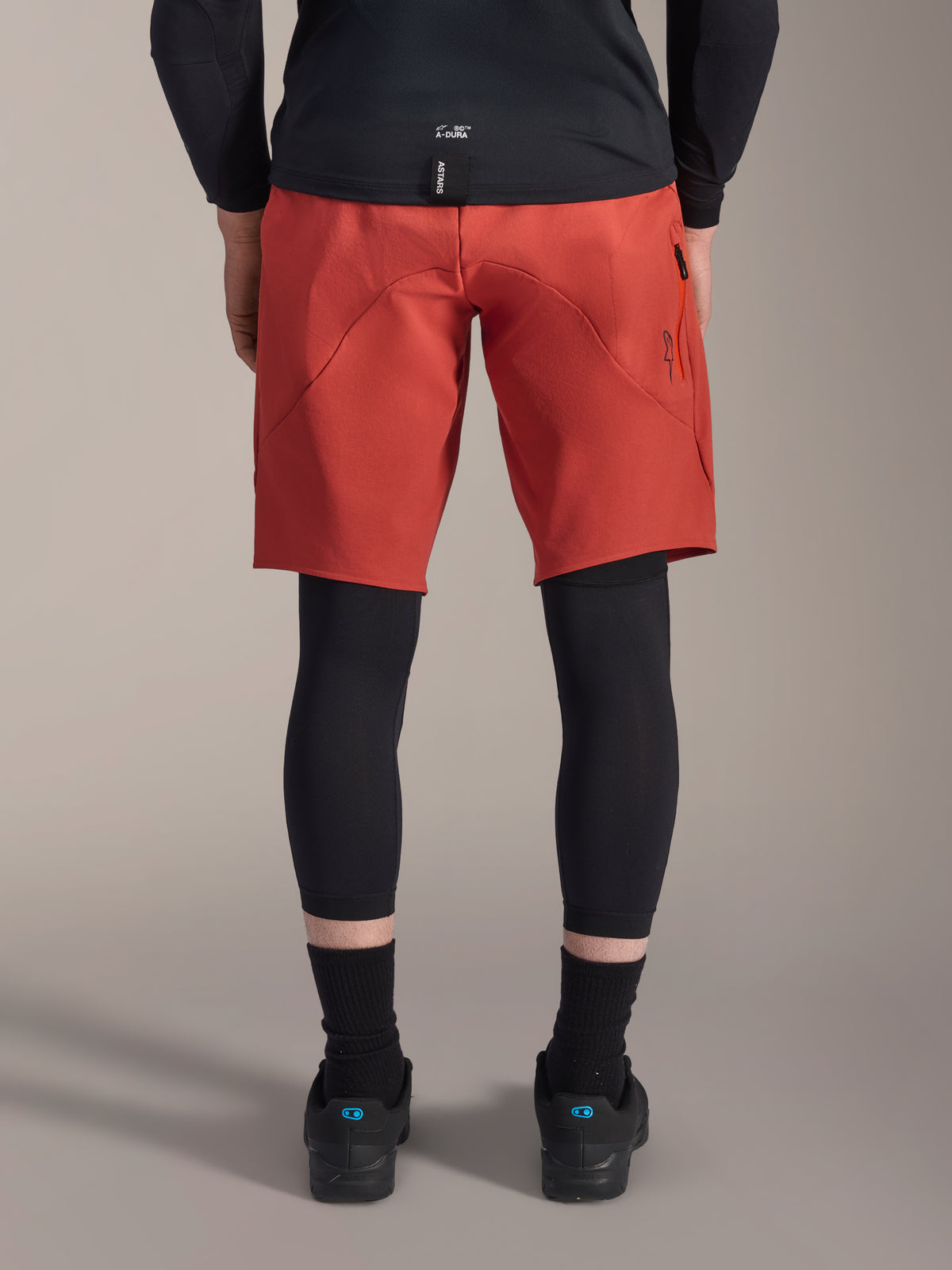 Joelheira Alpinestars A-Motion Plasma, protetores de joelho de BTT pretos, vista traseira, usados por baixo de calções de BTT vermelhos com uma camisola preta e sapatos técnicos de ciclismo, equipamento de proteção para BTT