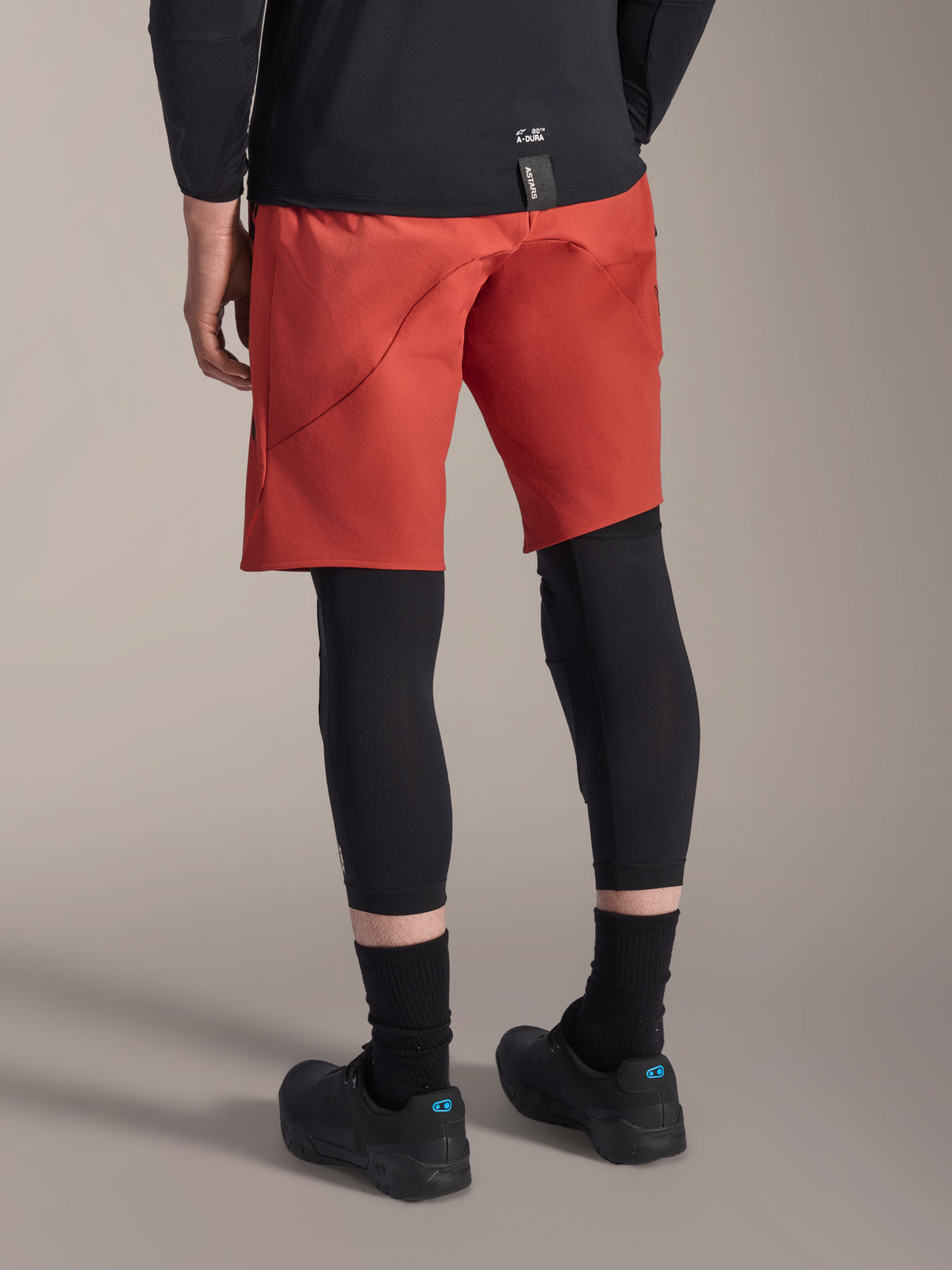 Joelheira Alpinestars A-Motion Plasma, protetor de joelho de MTB preto, usado com calções de BTT vermelhos e sapatilhas de ciclismo pretas, vista traseira mostrando o design slip-on de baixo perfil para ciclismo fora de estrada