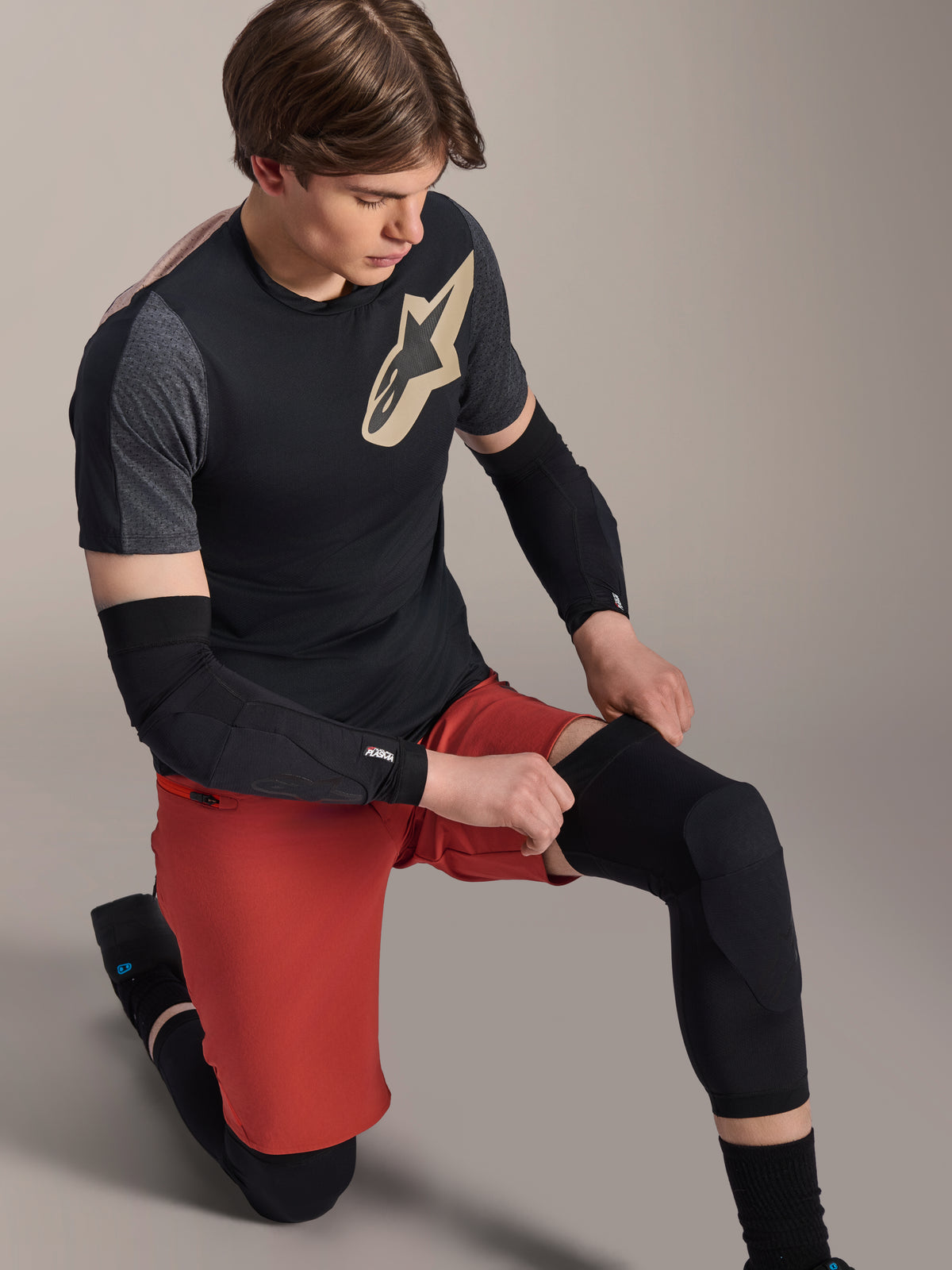 Alpinestars Joelheira A-Motion Plasma, protetor de joelho para MTB, preto, usado por um ciclista numa posição ajoelhada, combinado com uma t-shirt preta e cinzenta com logótipo dourado, calções vermelhos e cotoveleiras pretas, equipamento de proteção profissional para mountain bike