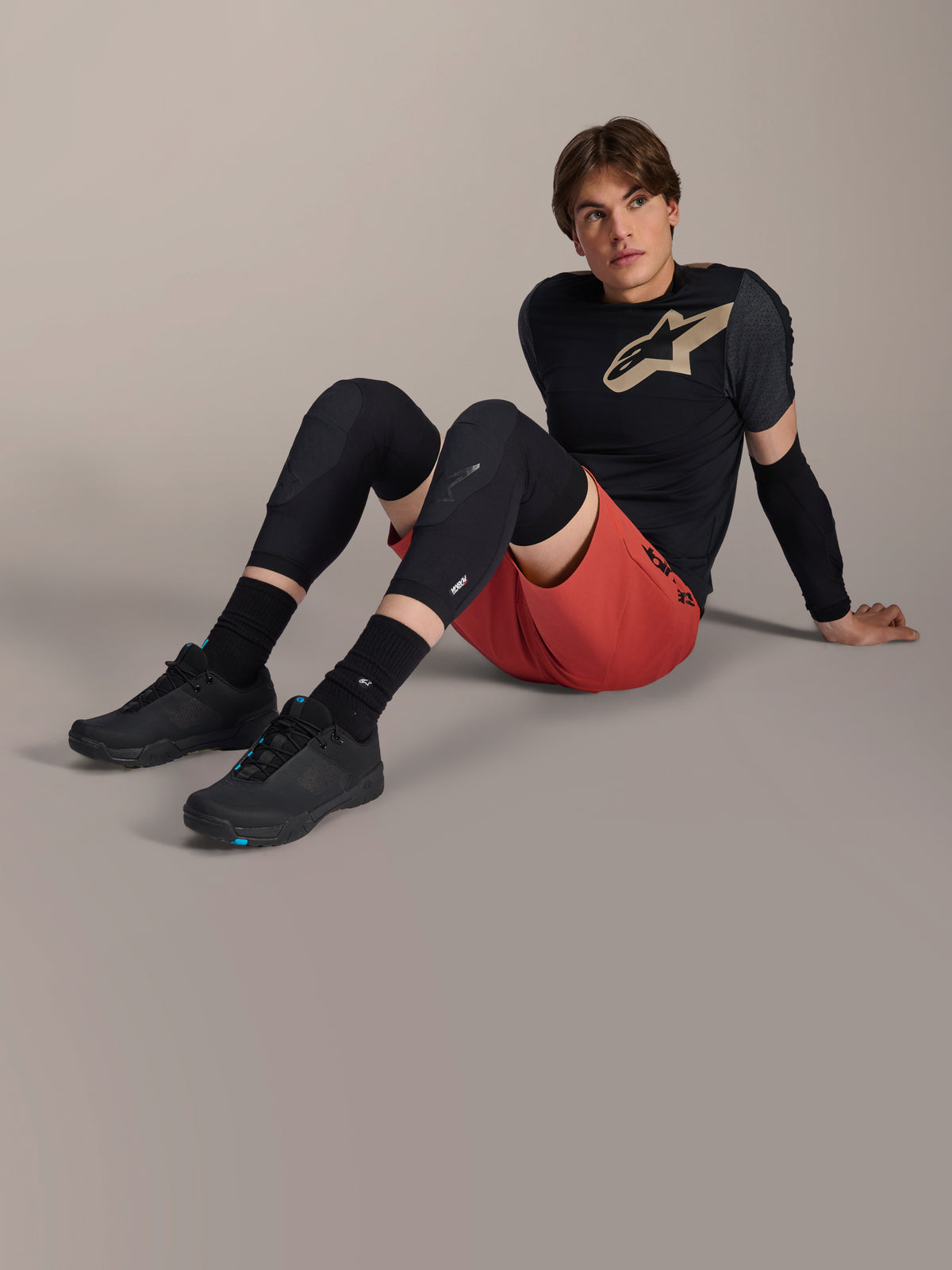 Alpinestars A-Motion Plasma Knee Sleeve, joelheiras pretas para BTT, usadas por um modelo masculino sentado com equipamento de ciclismo completo, calções vermelhos e t-shirt técnica preta, realçando o design protetor de baixo perfil