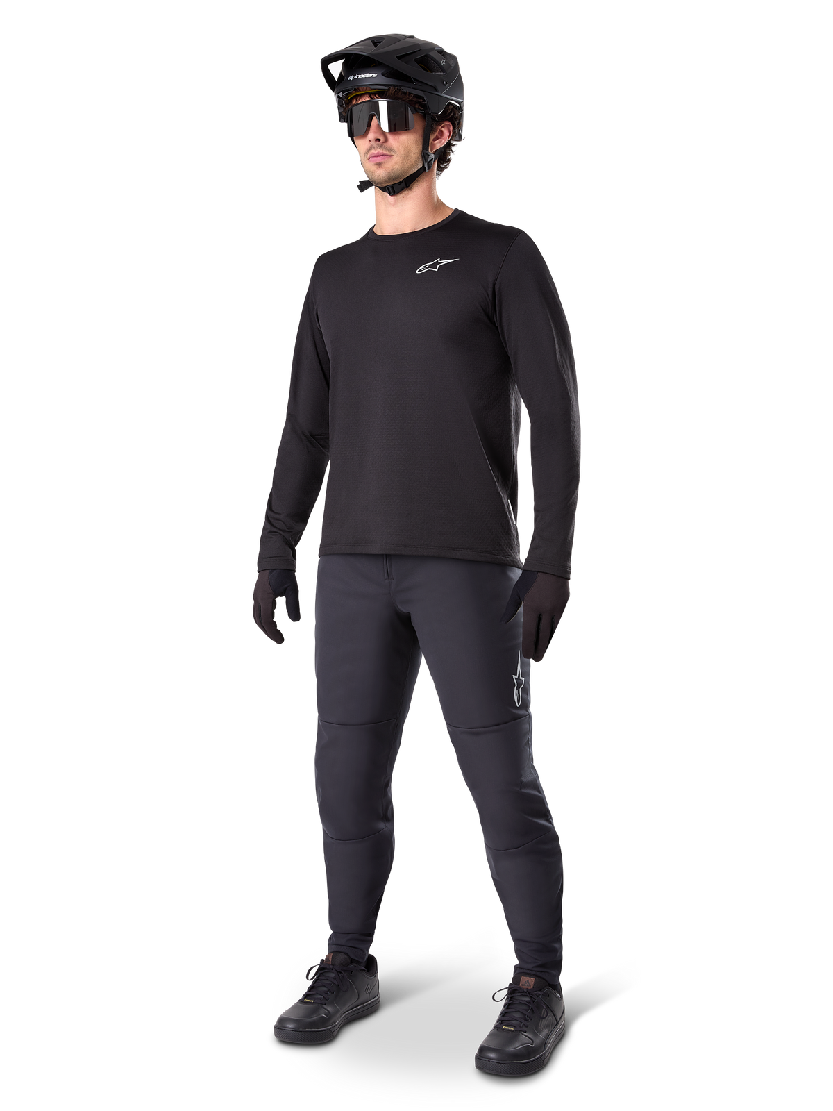 Ciclista de BTT a usar Calças Térmicas Alpinestars A-Dura, pretas, combinadas com uma camisola de manga comprida preta, capacete preto e óculos de sol, apresentando o logótipo prateado da Alpinestars na coxa esquerda, concebidas para a prática de trilhos em climas frios