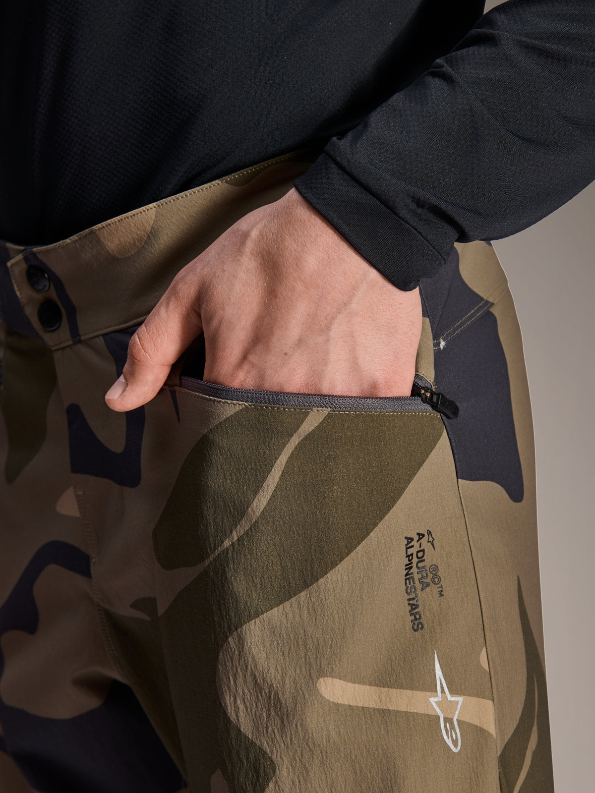 Pormenor das calças Alpinestars A-Dura Camo, calças de BTT com padrão de camuflagem castanha, apresentando uma mão num bolso com fecho, fecho de cintura com botões de pressão duplos e logótipo Alpinestars na coxa.