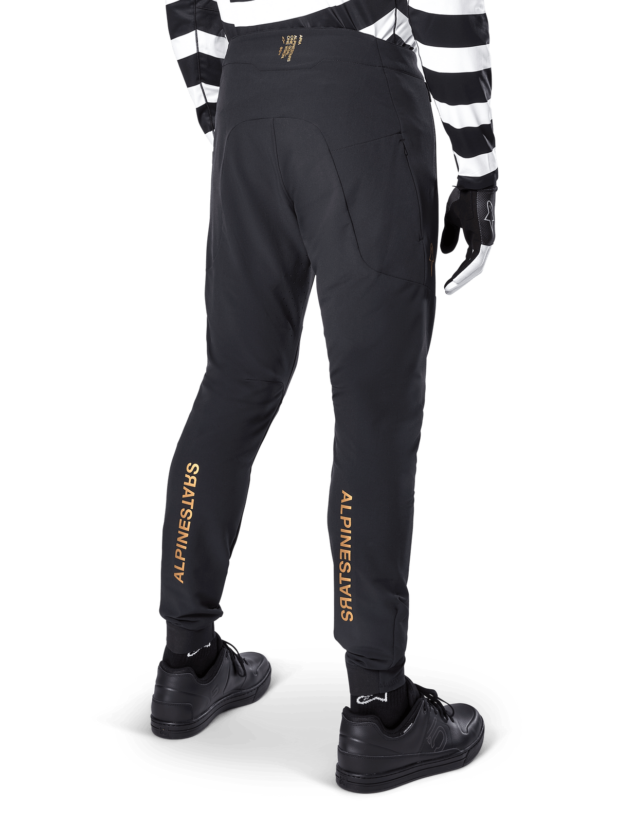 Alpinestars A-Aria Elite Pants, calças de BTT pretas, vista traseira, com logótipo Alpinestars vertical em dourado na parte de trás das pernas, usadas com camisola às riscas pretas e brancas e sapatos pretos, vestuário de performance para ciclismo de montanha