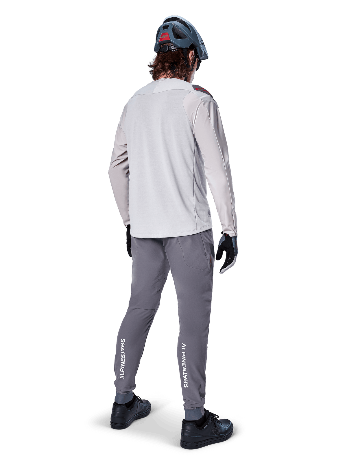 Calças Alpinestars A-Aria Elite, calças de ciclismo MTB, cinzento escuro, vista traseira de um ciclista com o equipamento completo, com logótipo Alpinestars em branco na parte inferior das pernas, ajuste de performance com perfurações a laser, combinadas com uma camisola de manga comprida cinzento claro e capacete de MTB cinzento