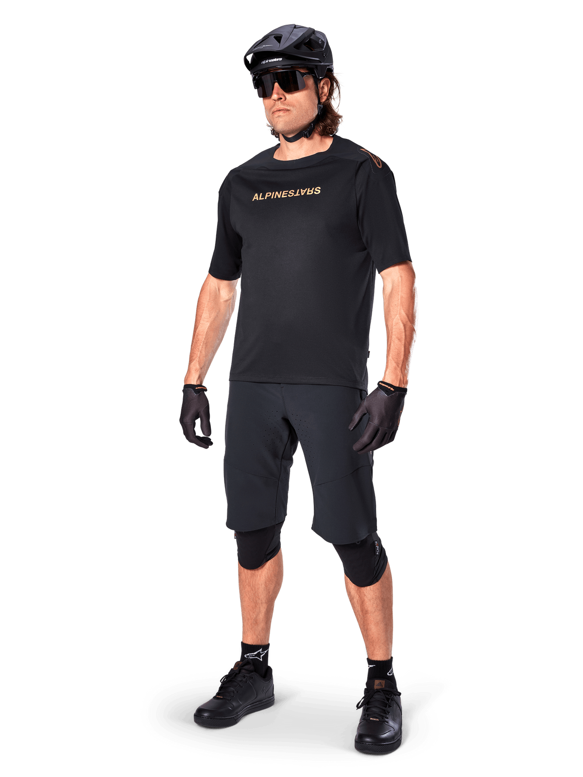 Ciclista a usar Calções Alpinestars A-Aria Elite, calções de BTT pretos, combinados com uma camisola preta com logótipo dourado, capacete preto, óculos de sol e joelheiras, equipamento de performance completo para ciclismo fora de estrada
