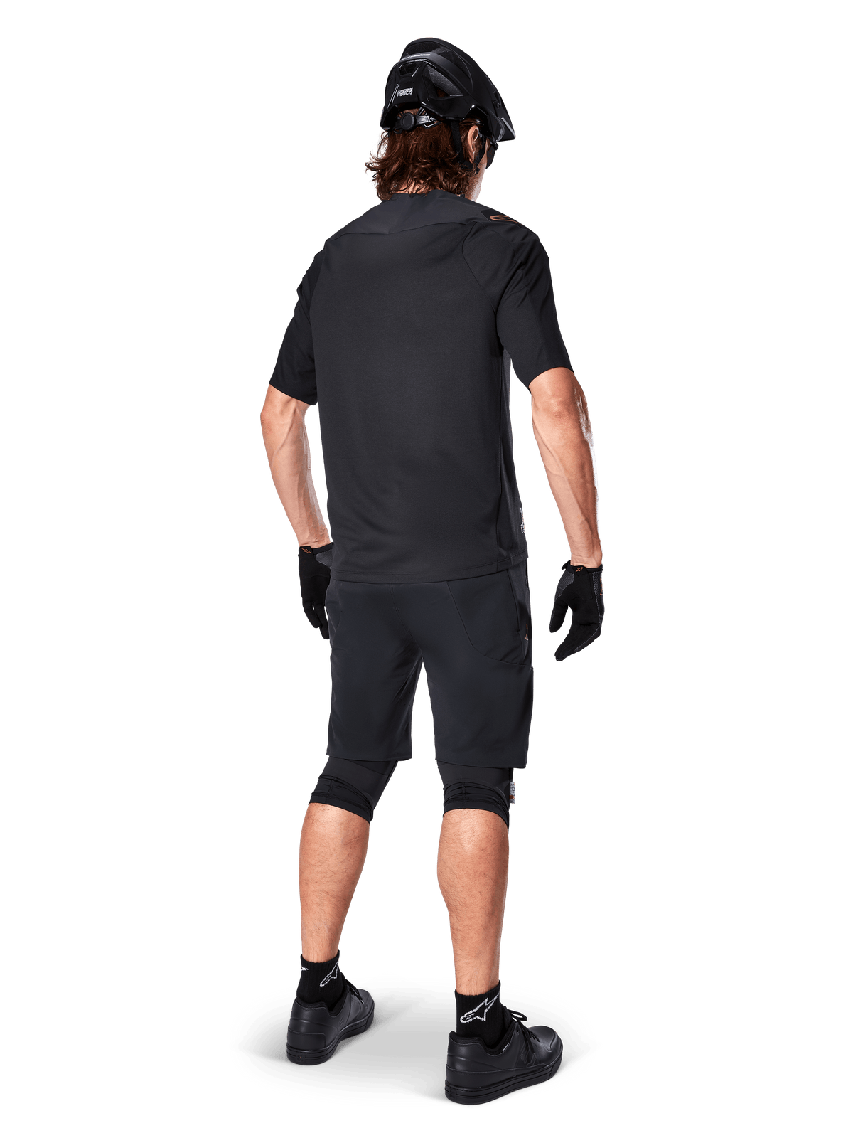 Vista traseira de um ciclista de montanha a usar calções Alpinestars A-Aria Elite em preto, combinados com uma camisola de manga curta preta a condizer, capacete preto, luvas e sapatos de BTT, mostrando um equipamento de performance completo para ciclismo fora de estrada.