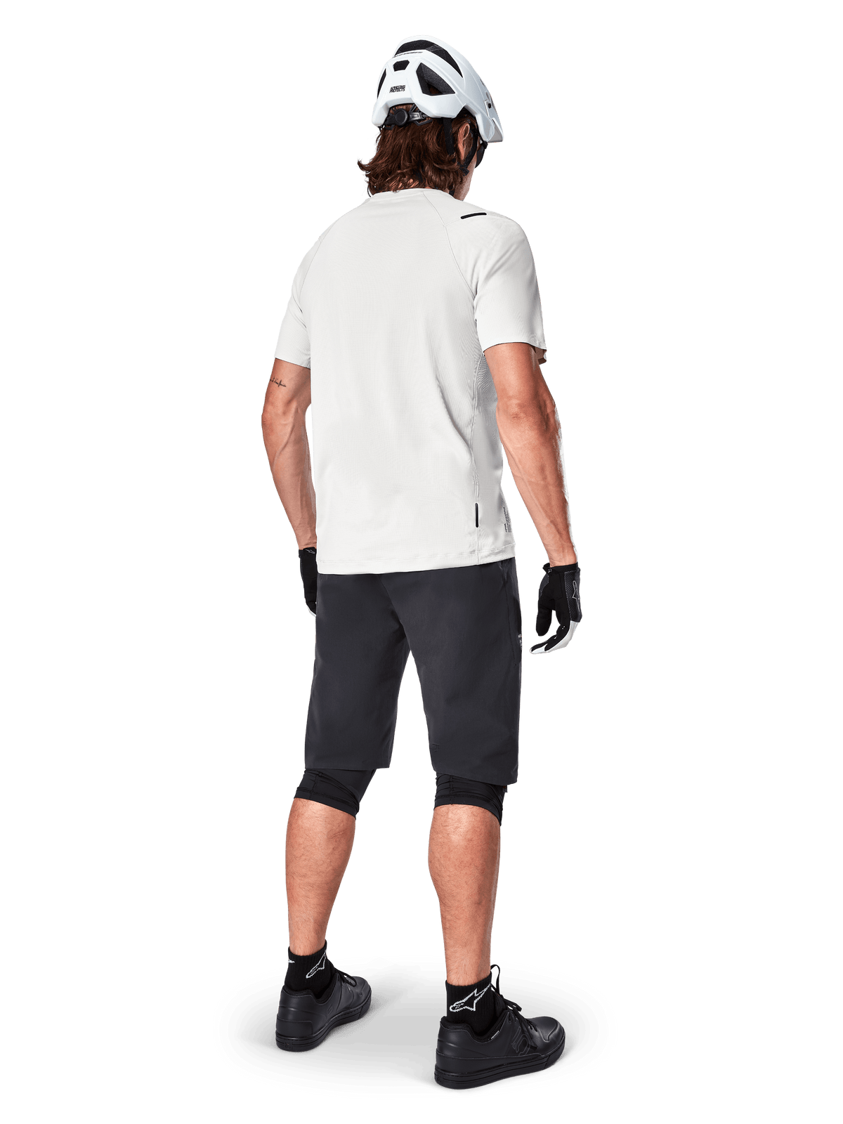 Ciclista de montanha visto de trás a usar calções Alpinestars A-Aria pretos, camisola de performance branca de manga curta, capacete branco, luvas pretas e sapatos de BTT pretos, equipamento de ciclismo para trilhos