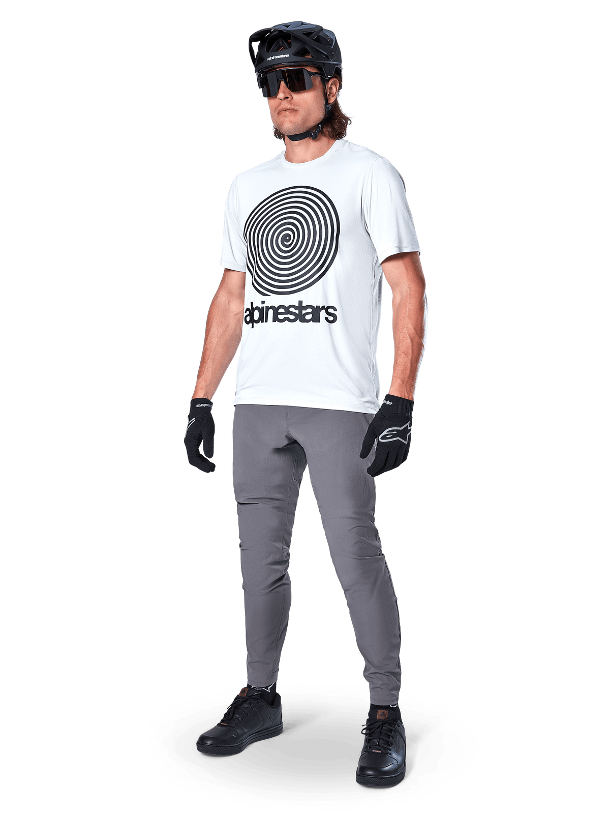Calças Alpinestars A-Dura, calças de MTB, cinza escuro, usadas com t-shirt branca com grafismo em espiral, capacete de MTB preto, óculos de sol e luvas pretas, tecido durável com elasticidade em 2 direções para ciclismo fora de estrada