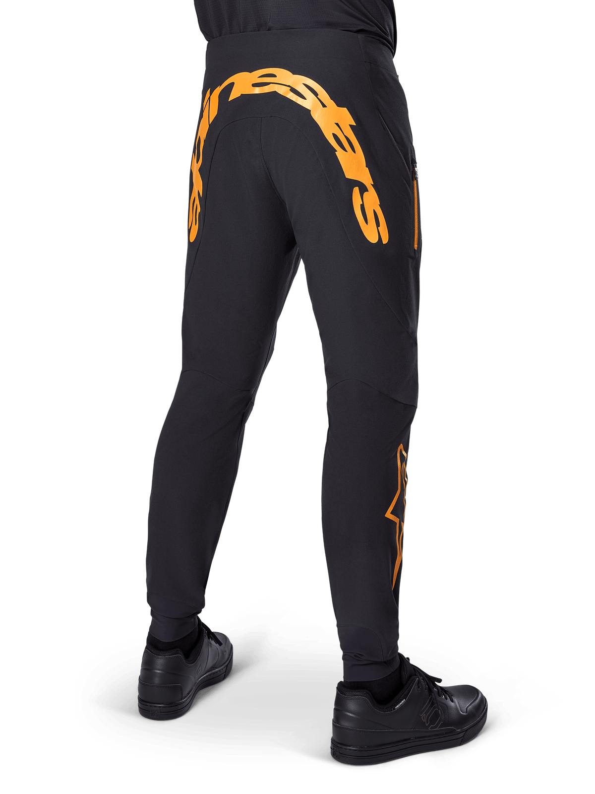 Calças Alpinestars A-Supra Race, calças de MTB pretas, vista traseira com grande logótipo Alpinestars cor de laranja na pala traseira, logótipo laranja na parte inferior da perna, ajuste de performance com bolso com fecho de correr, usadas com sapatos pretos