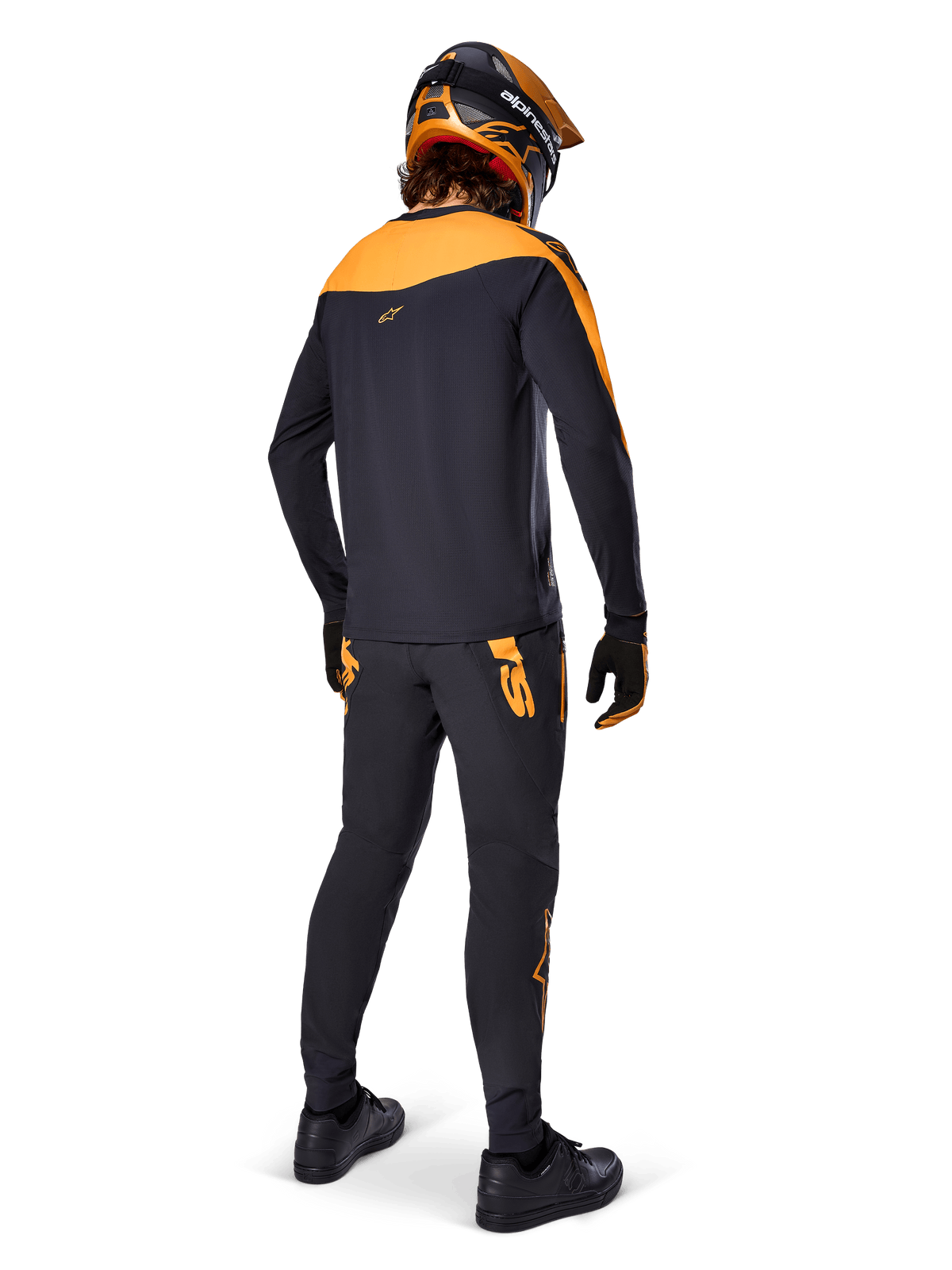 Calças A-Supra Race da Alpinestars, calças de corrida de BTT, pretas com logótipos laranja, vista posterior de um ciclista a usar camisola, capacete e luvas em preto e laranja coordenados, realçando o logótipo inspirado em MX na pala traseira e o ajuste de performance para trilhos técnicos.