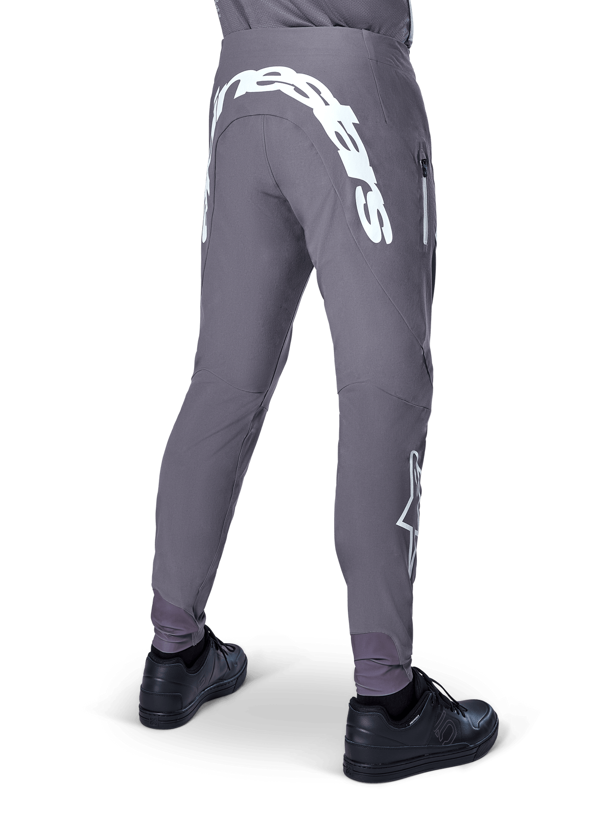 Calças Alpinestars A-Supra Race, calças de mountain bike, cinzento escuro, vista traseira com sapatos pretos, com uma grande inscrição Alpinestars branca na pala traseira e logótipo de estrela na perna, bolso com fecho de correr, tecido elástico de performance para corridas de BTT