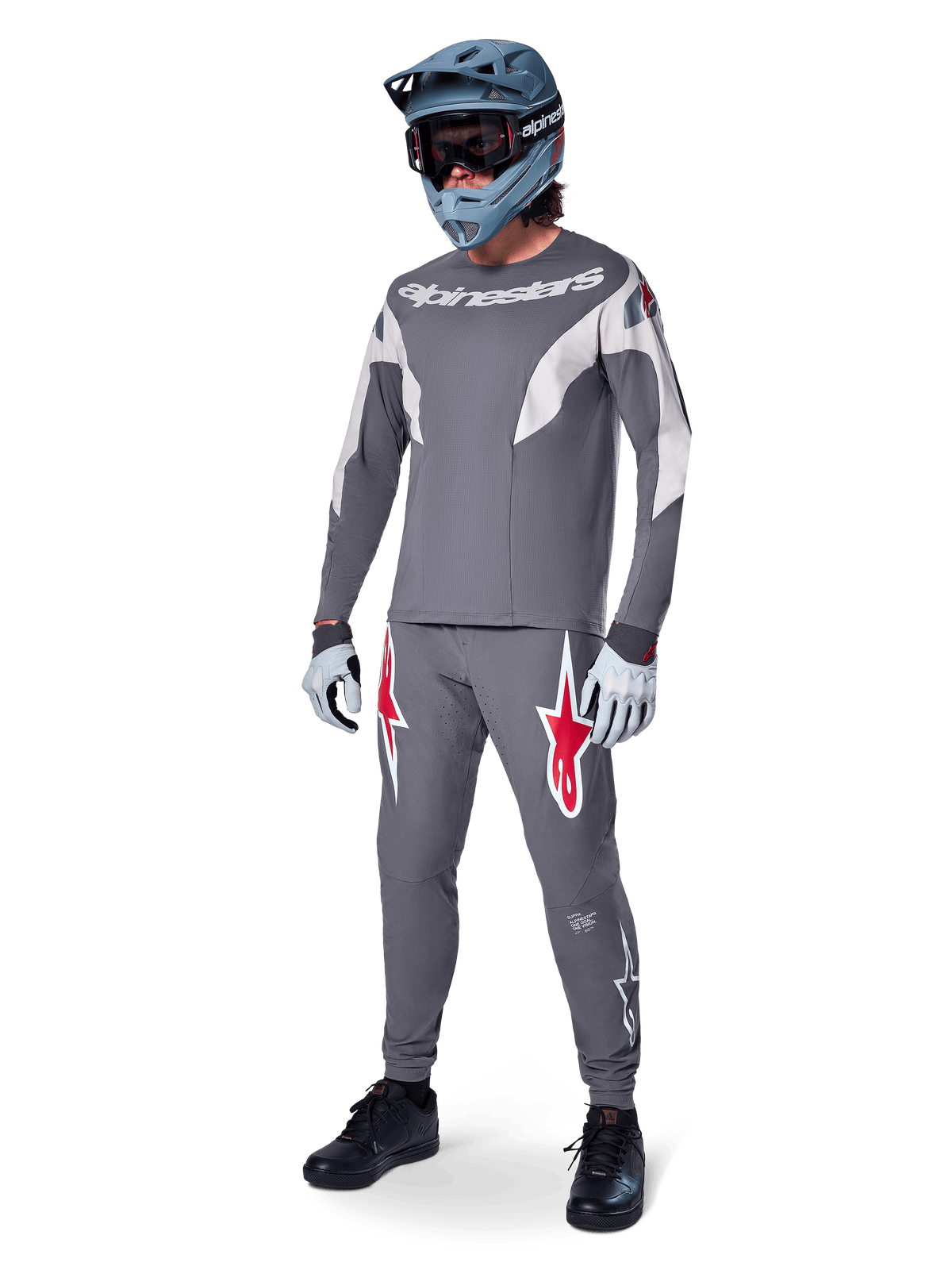 Alpinestars A-Supra Race Pants em cinzento escuro, com logótipos vermelhos e brancos destacados nas coxas, usadas por um ciclista como parte de um kit completo de corrida de BTT com camisola cinzenta a condizer, capacete integral azul-acinzentado e luvas cinzentas claras, equipamento de alta performance para corridas de MTB