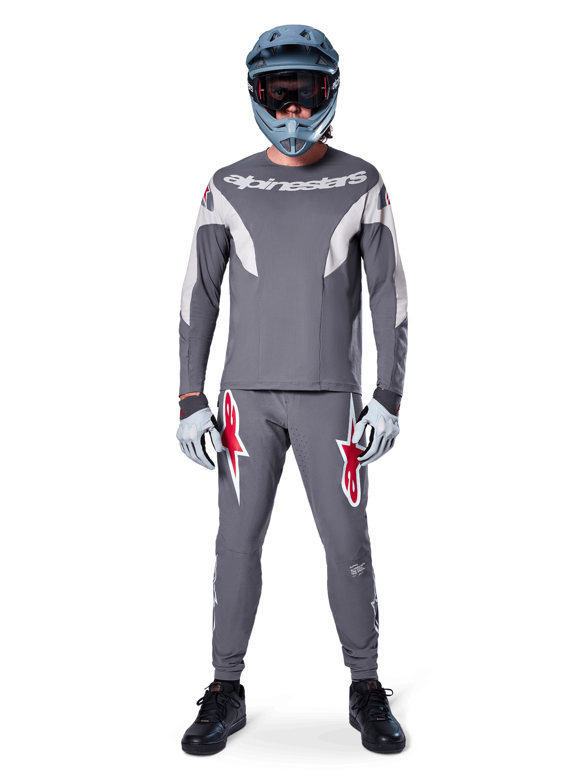 Ciclista de MTB a usar calças Alpinestars A-Supra Race em cinzento escuro, com logótipos vermelhos destacados nas coxas, combinadas com uma camisola cinzenta a condizer, capacete integral azul e sapatilhas pretas para um kit completo de corrida todo-o-terreno