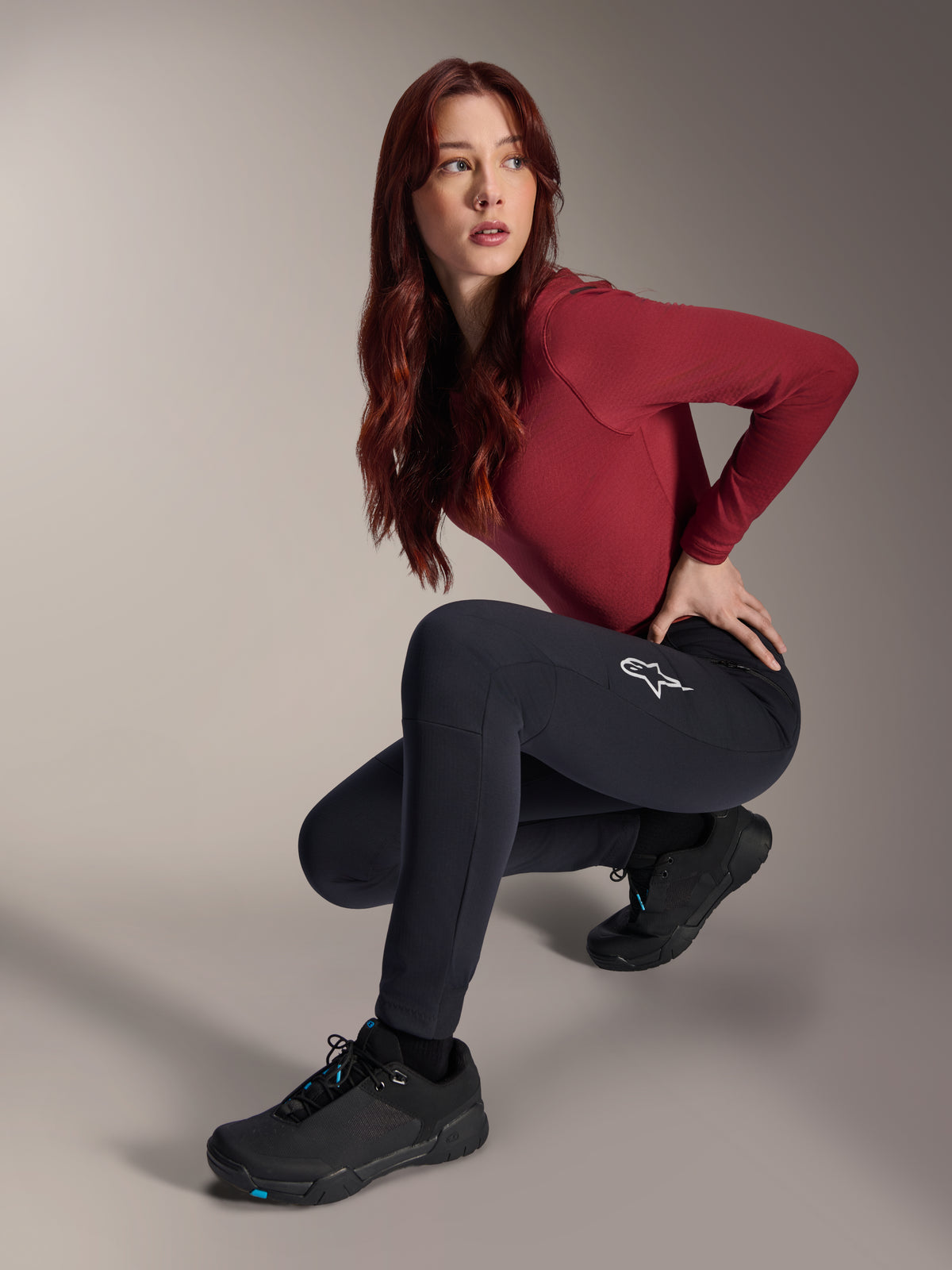 Calças Térmicas Alpinestars Stella A-Dura, calças de MTB para mulher, pretas, usadas por uma modelo numa pose agachada com uma camisola de manga comprida bordeaux e sapatos de ciclismo pretos, apresentando um logótipo de estrela branca na coxa, construção durável e quente para ciclismo em clima frio