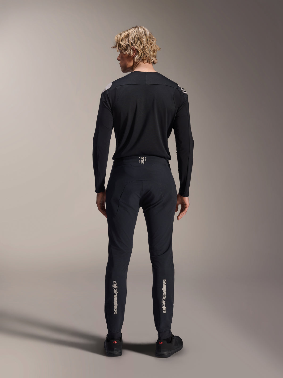 Calças Alpinestars A-Aria Elite, Black Titanium, vista traseira num modelo masculino, com a marca Alpinestars em prateado na parte inferior das pernas, calças técnicas de MTB com um ajuste de performance e detalhe no cós que absorve a humidade.
