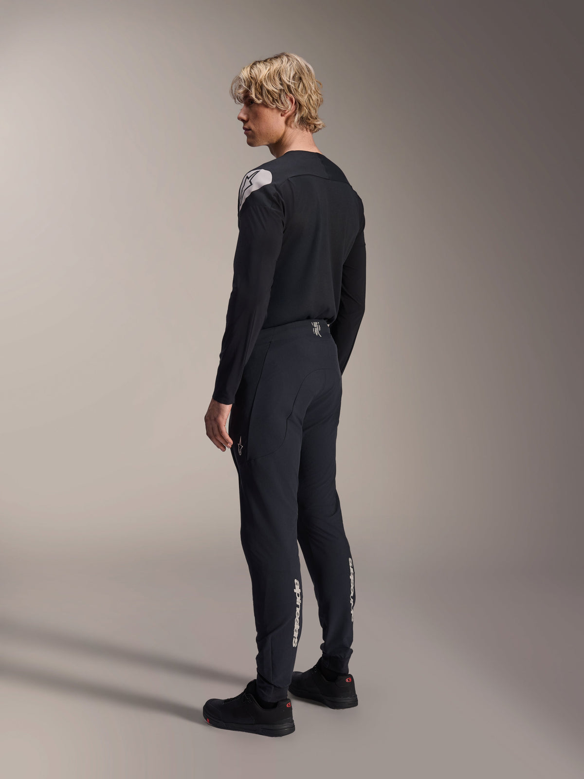 Alpinestars A-Aria Polartec Elite Jersey - Manga Comprida, preta, vista de perfil lateral traseiro num modelo, com logótipo branco no ombro, combinada com calças de ciclismo pretas a condizer para um conjunto de MTB profissional