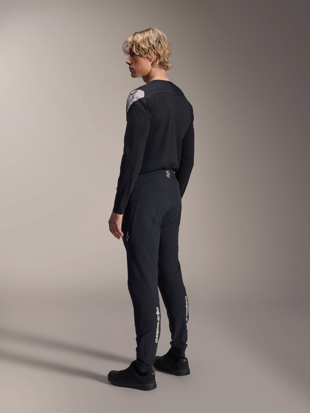 Calças Alpinestars A-Aria Elite, calças de mountain bike, Black Titanium, vista traseira de três quartos no modelo, com o logótipo Alpinestars branco na parte inferior das pernas, corte de performance com punhos elásticos, combinadas com uma camisola de manga comprida preta a condizer.