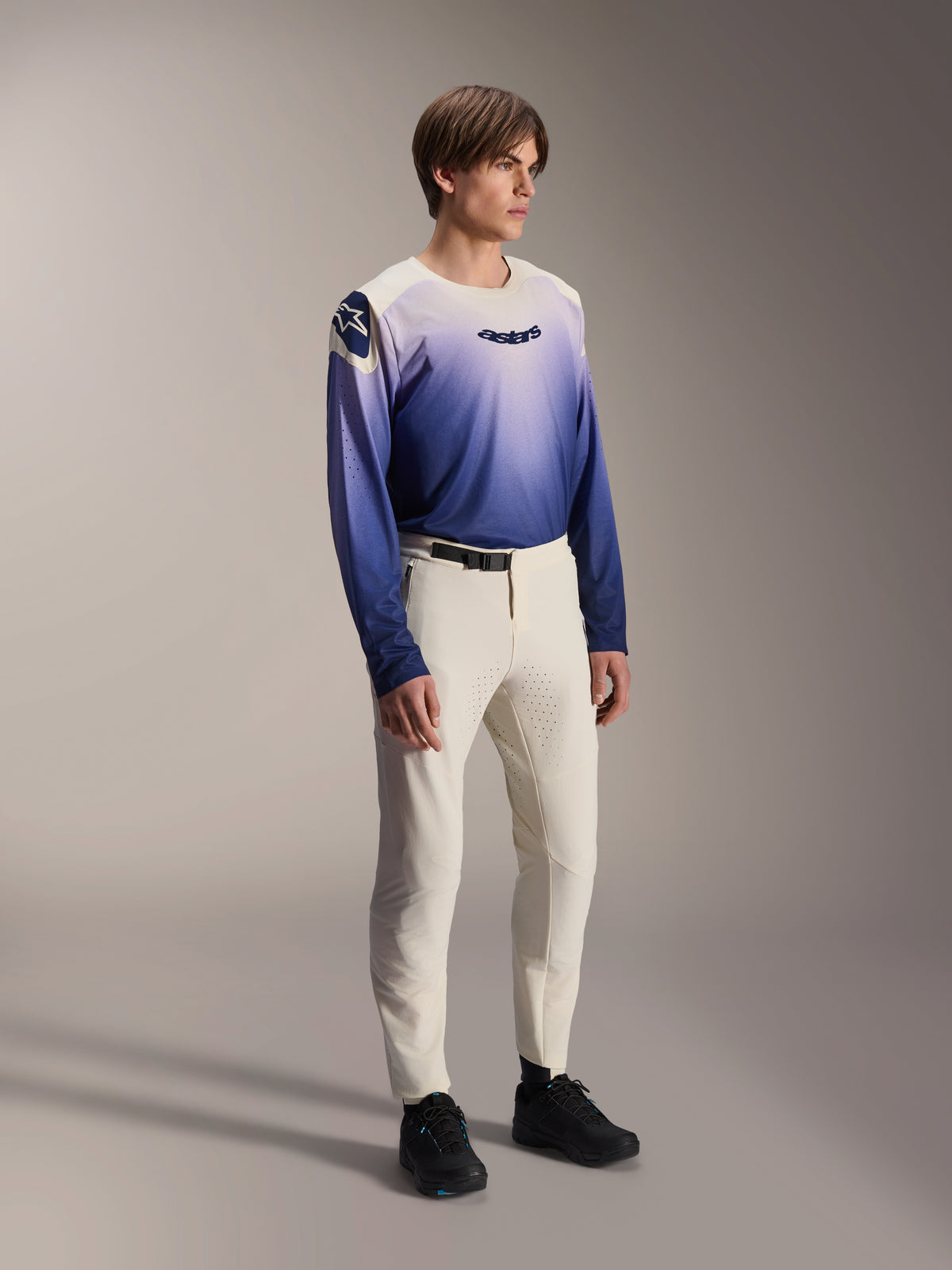 Model wearing Alpinestars A-Aria Elite Pants in Ecru, off-white mountain bike pants with laser-cut ventilation and a black waist buckle, paired with a blue and purple gradient long-sleeve jersey, performance-driven cycling gear for off-road trails