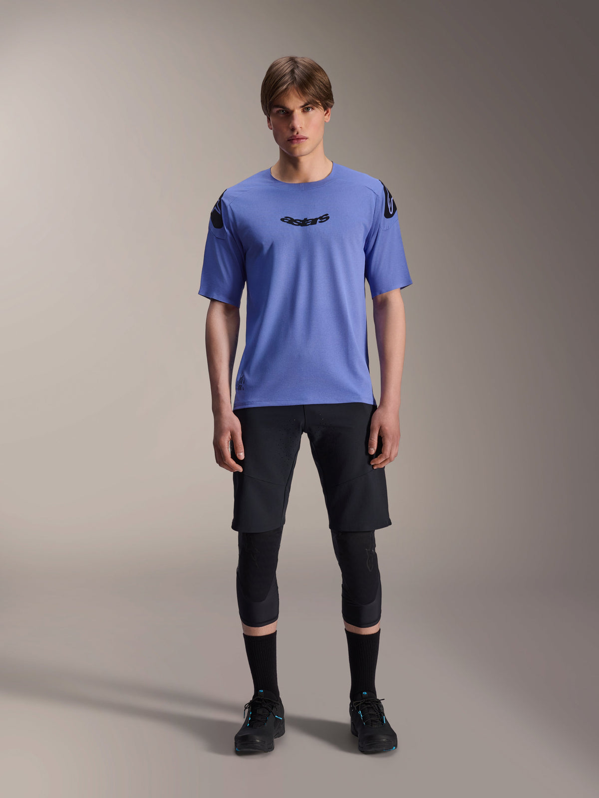 Modelo masculino a usar camisola de manga curta Alpinestars A-Aria Polartec Elite em Baja Blue, logótipo astars preto no peito, combinada com calções de BTT pretos, joelheiras e sapatos de ciclismo pretos, fotografia de estúdio para vestuário de mountain bike