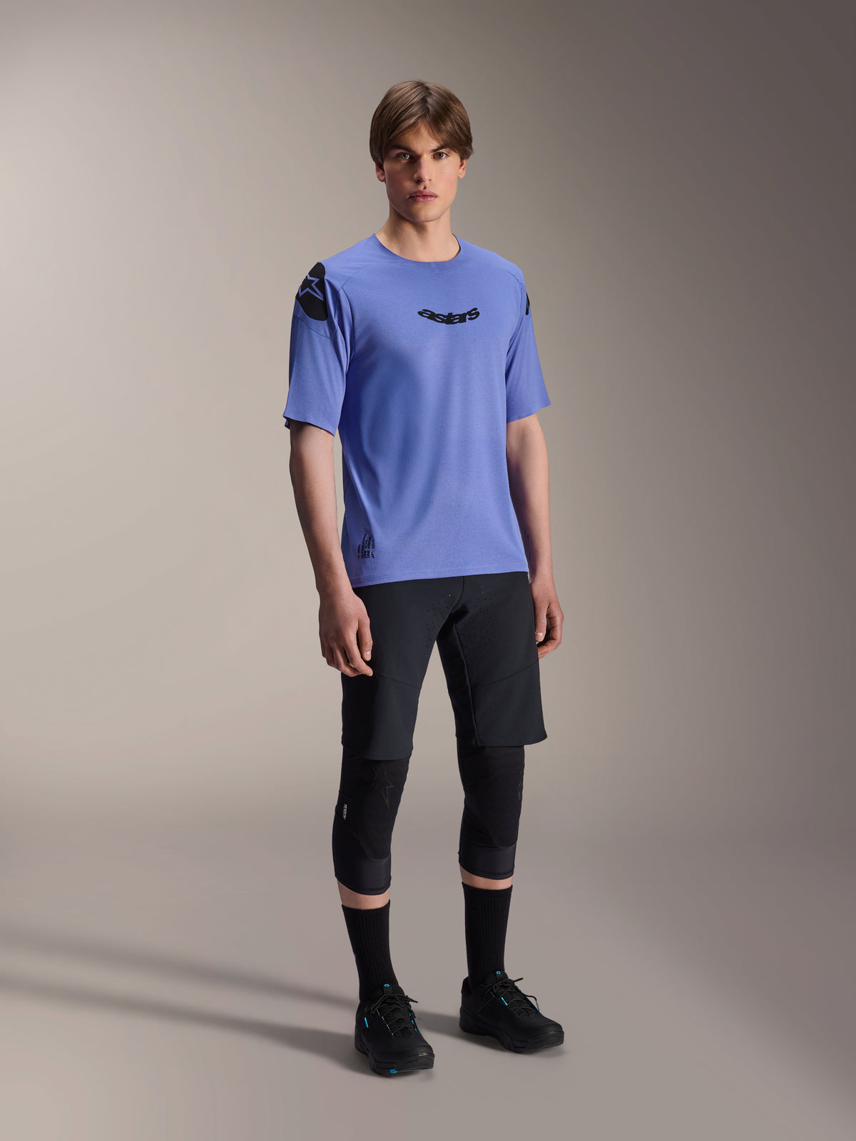 Modelo masculino a vestir calções Alpinestars A-Aria Lite em preto, combinados com uma camisola de manga curta A-Aria Polartec Elite azul, joelheiras pretas e sapatos de ciclismo pretos, vestuário de performance para BTT em trilhos.