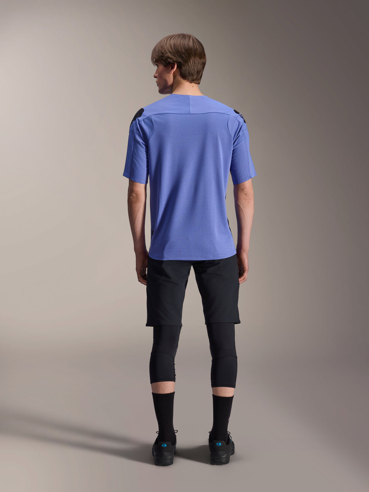 Male model from the back wearing Alpinestars mountain bike gear, light blue A-Aria Polartec Elite short sleeve jersey, black A-Aria Elite Shorts, black knee protectors, black socks, and black shoes, coordinated performance outfit for MTB.