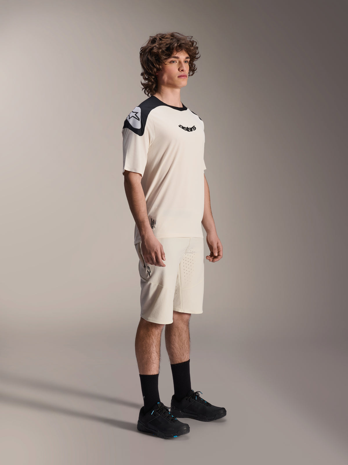 Modelo masculino a usar calções Alpinestars A-Aria Elite em Ecru, calções técnicos de BTT com ventilação cortada a laser, combinados com uma camisola de manga curta em creme e preto a condizer e sapatos pretos, vestuário de ciclismo profissional