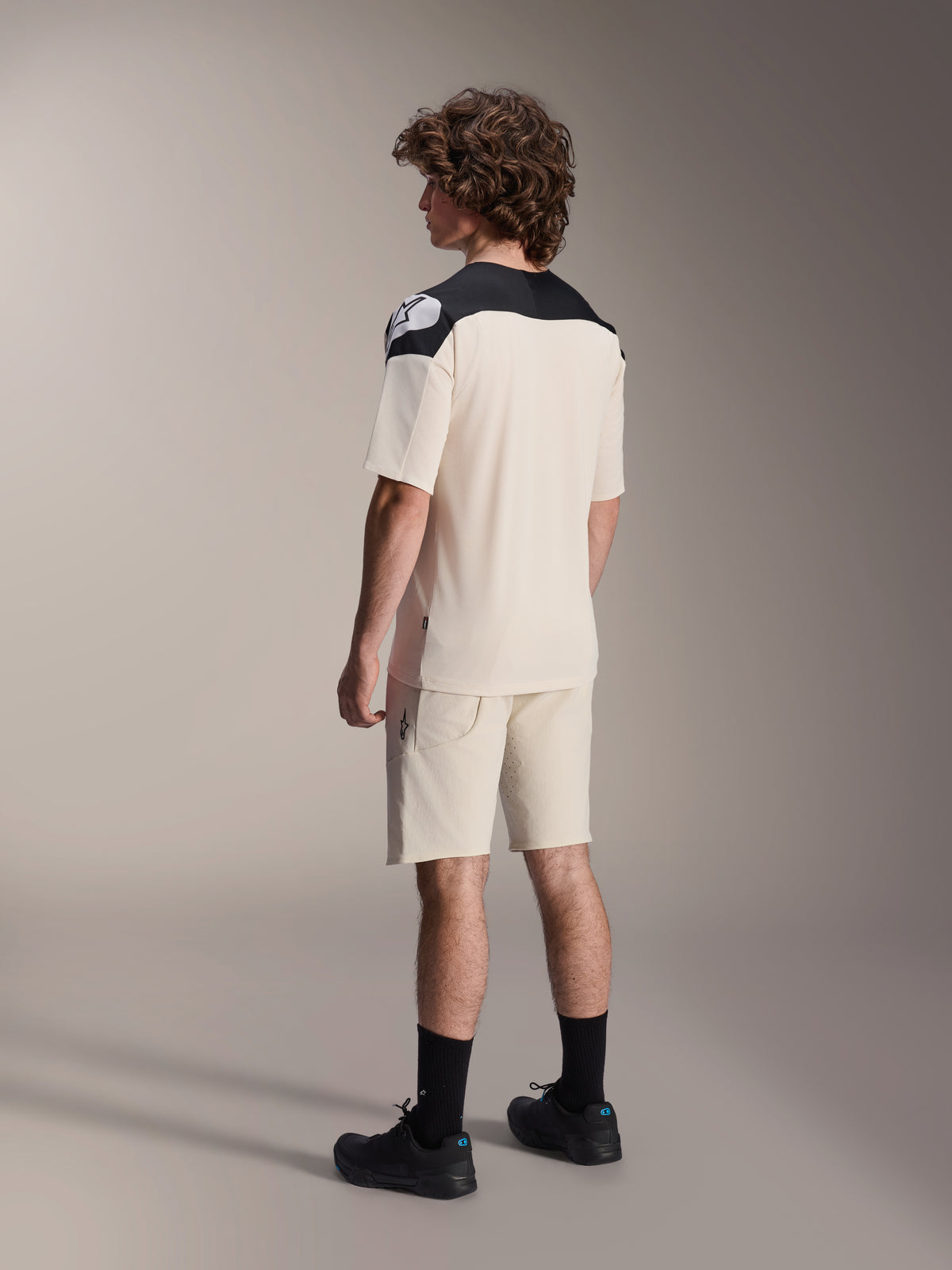 Calções Alpinestars A-Aria Elite e camisola a condizer, vestuário de Mountain Bike, cor cru com detalhes pretos, modelo masculino visto de costas com meias e sapatos pretos, equipamento de ciclismo de alta performance para todo-o-terreno